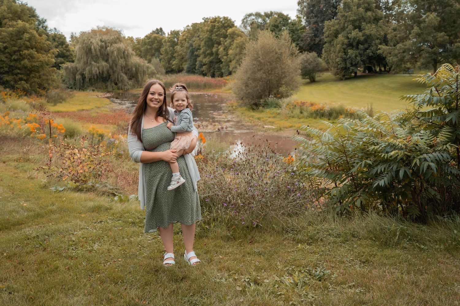 A woman in a sage green dress holding a child stands in a scenic autumn meadow beside a stream.
