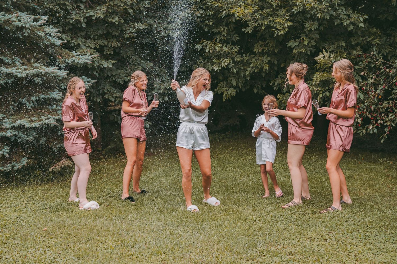 Wedding party members in pink outfits dance in line on grassy outdoor area.