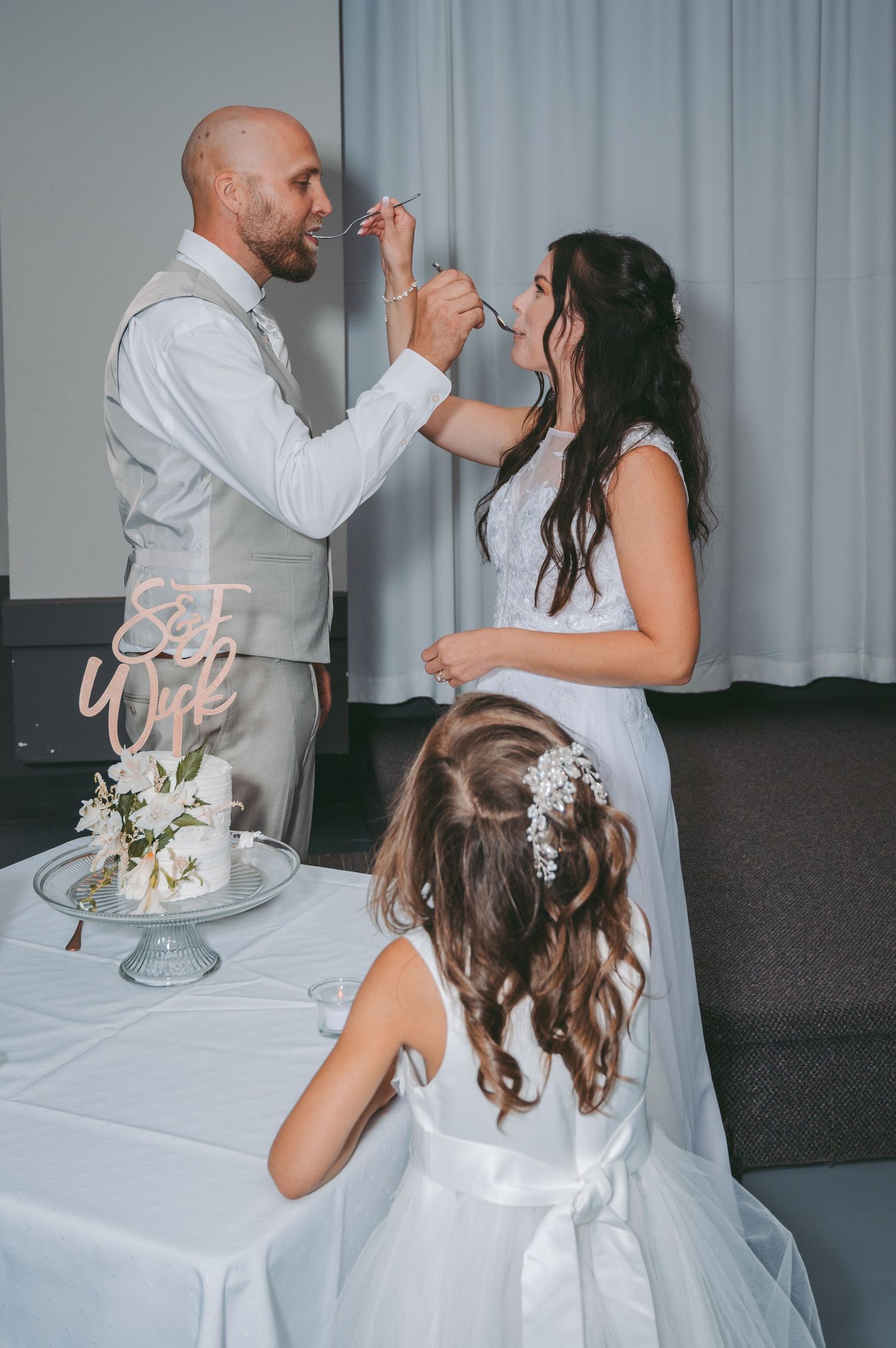 Couple shares a playful cake-tasting moment at their wedding reception while a child watches from below.