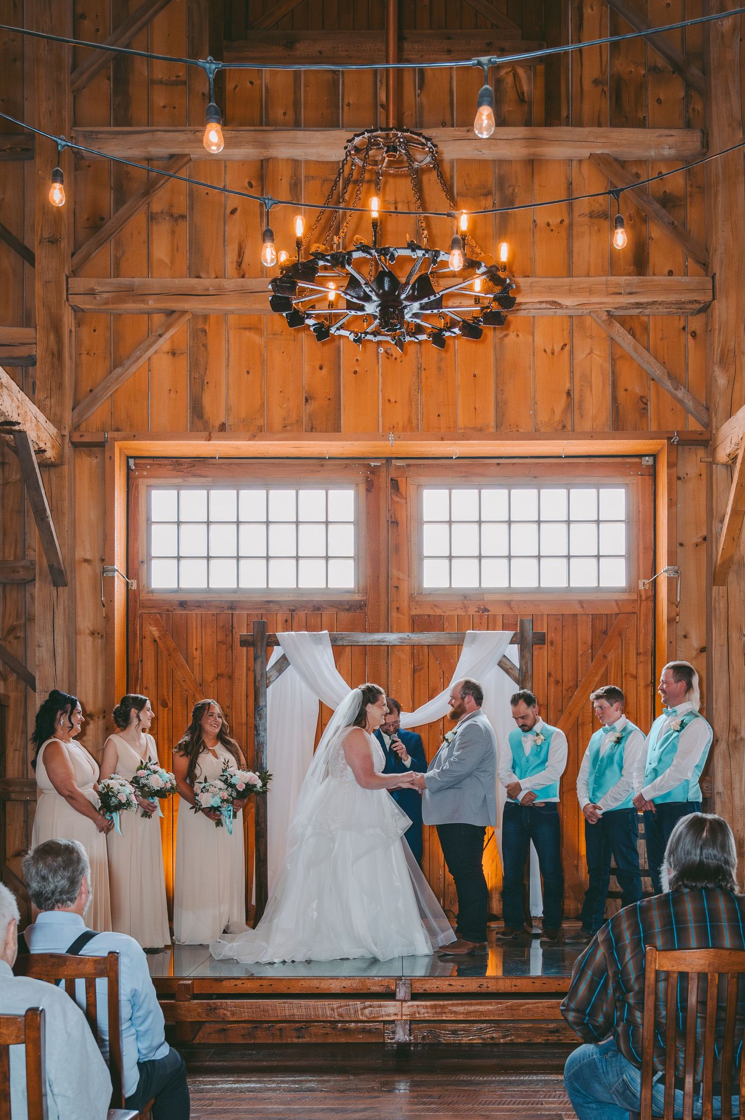 Wedding ceremony in a rustic barn with wooden beams, chandelier, and natural light streaming through windows.