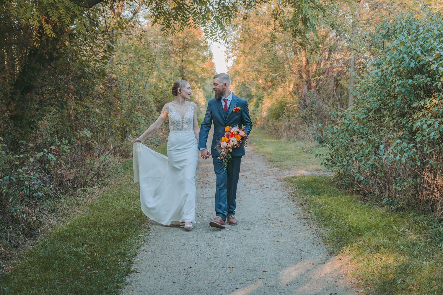 A wedding couple walks hand in hand down a nature path during golden hour.