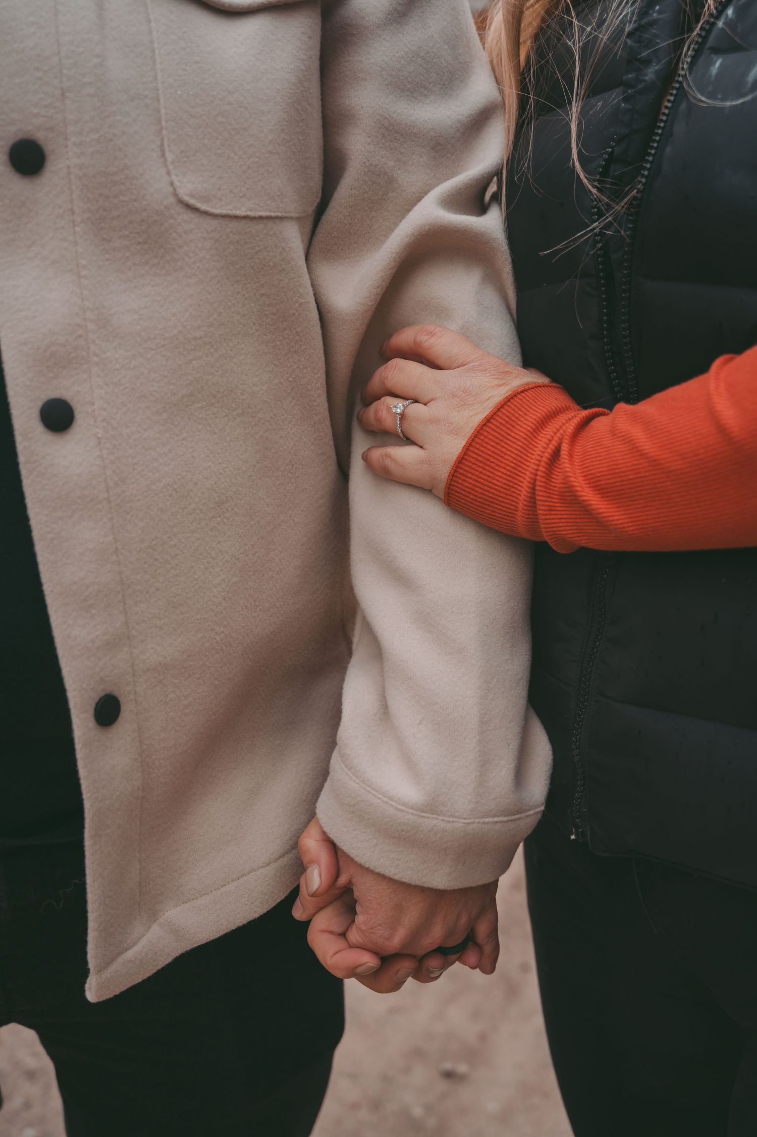 A close-up of clasped hands and a beige coat showing an engagement ring.
