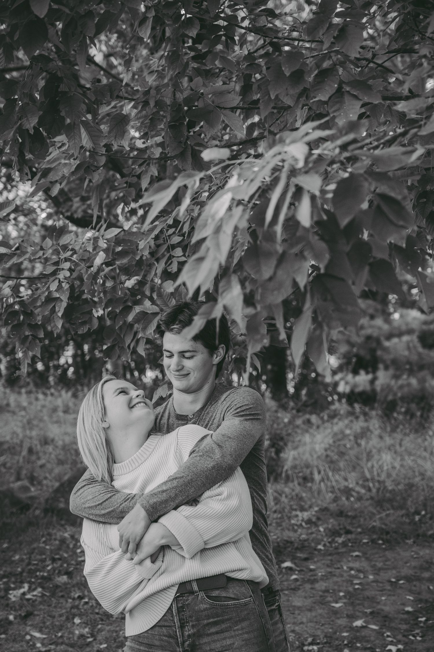 A tender black and white moment captured of a couple embracing under tree branches.