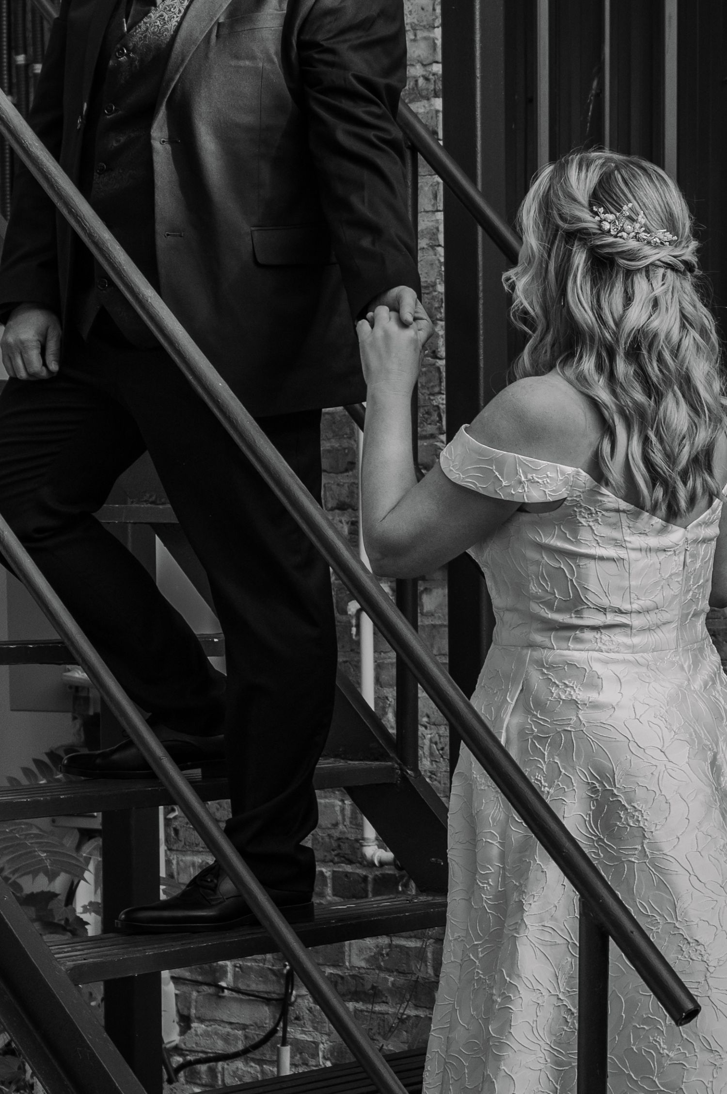 A bride in a white lace wedding dress walks up a dark metal staircase with exposed brick walls.