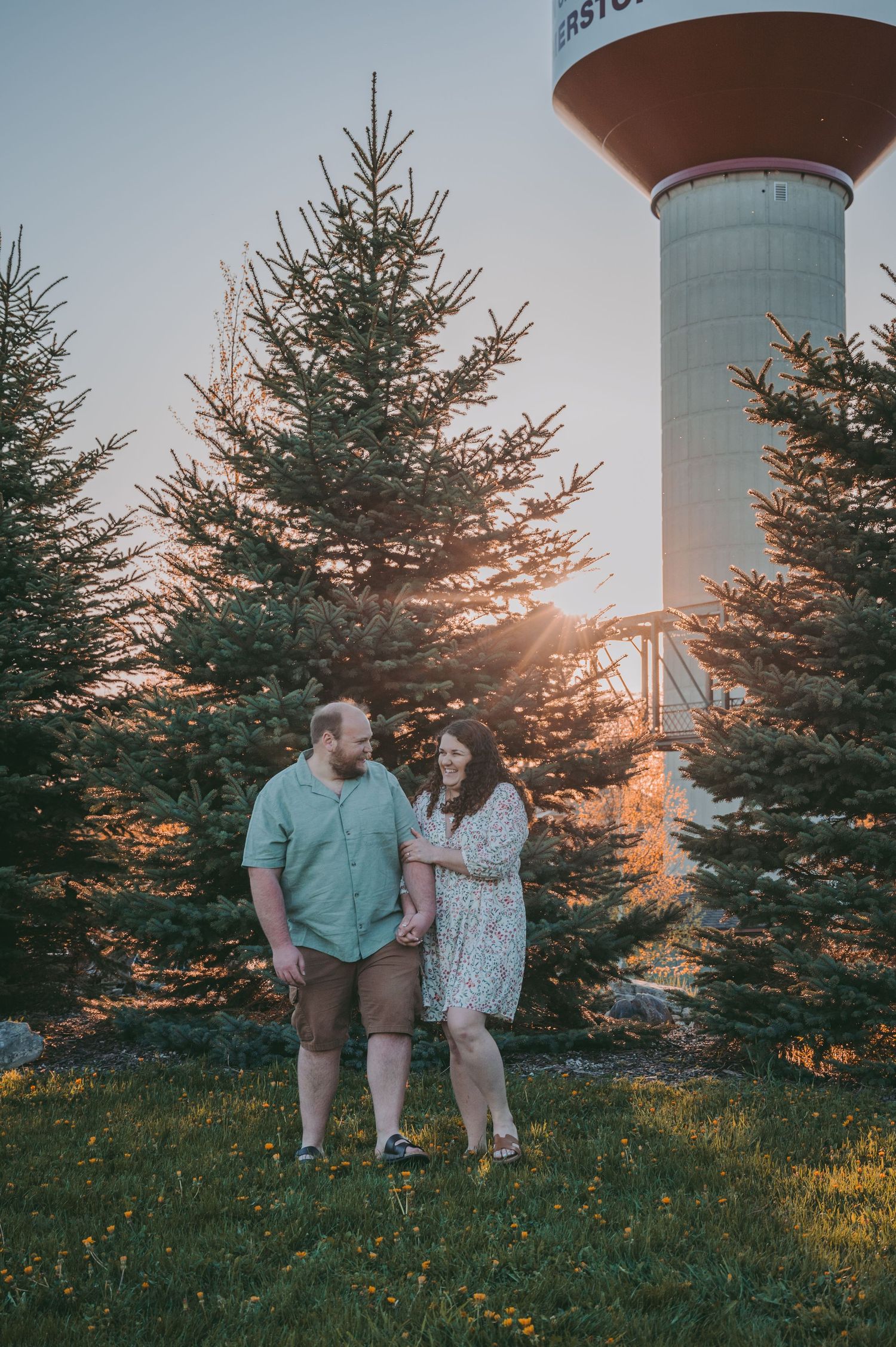 A couple poses together at sunset near evergreen trees and a water tower.