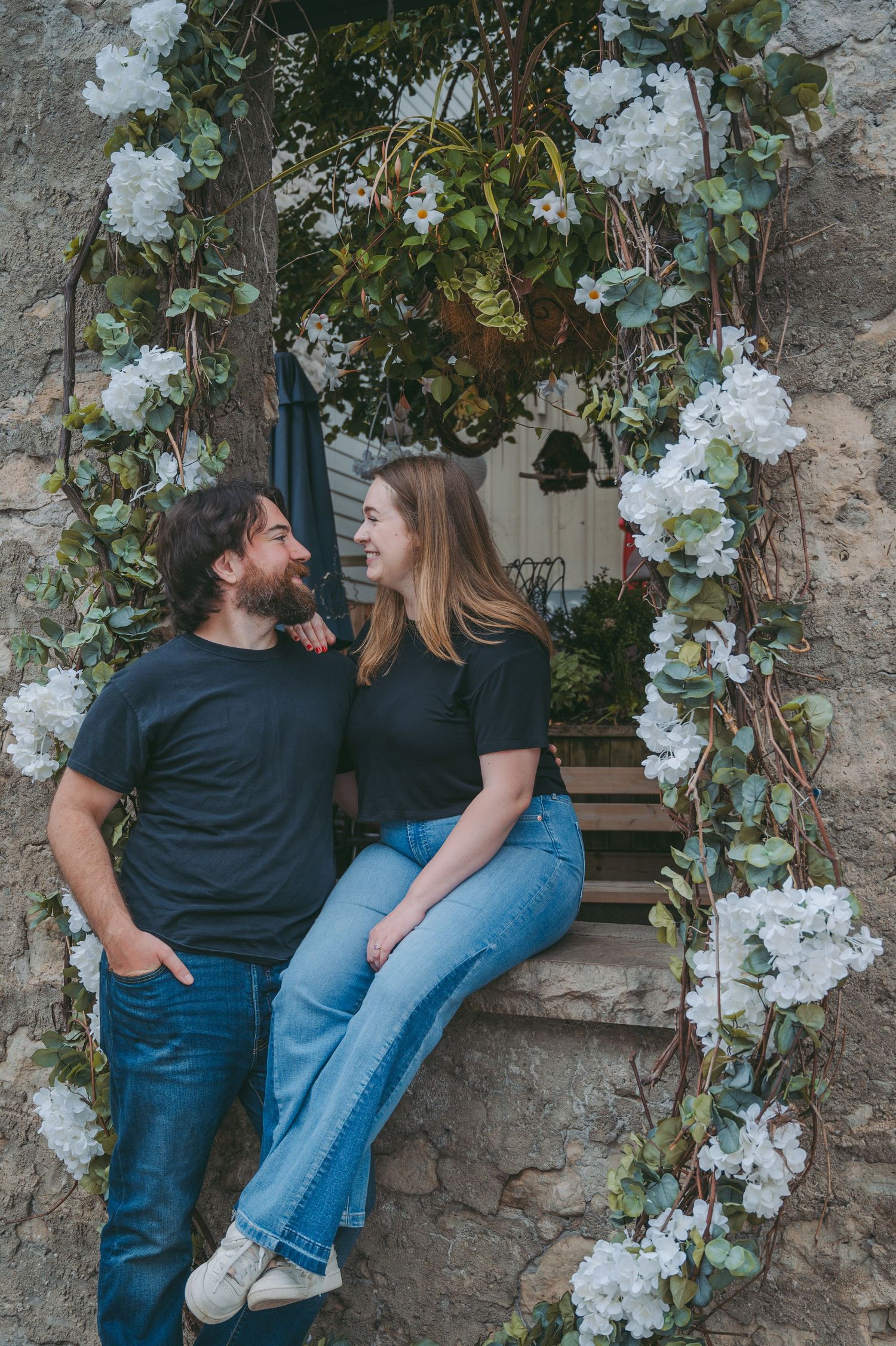 Couple sharing romantic moment by floral arch with white flowers and greenery.