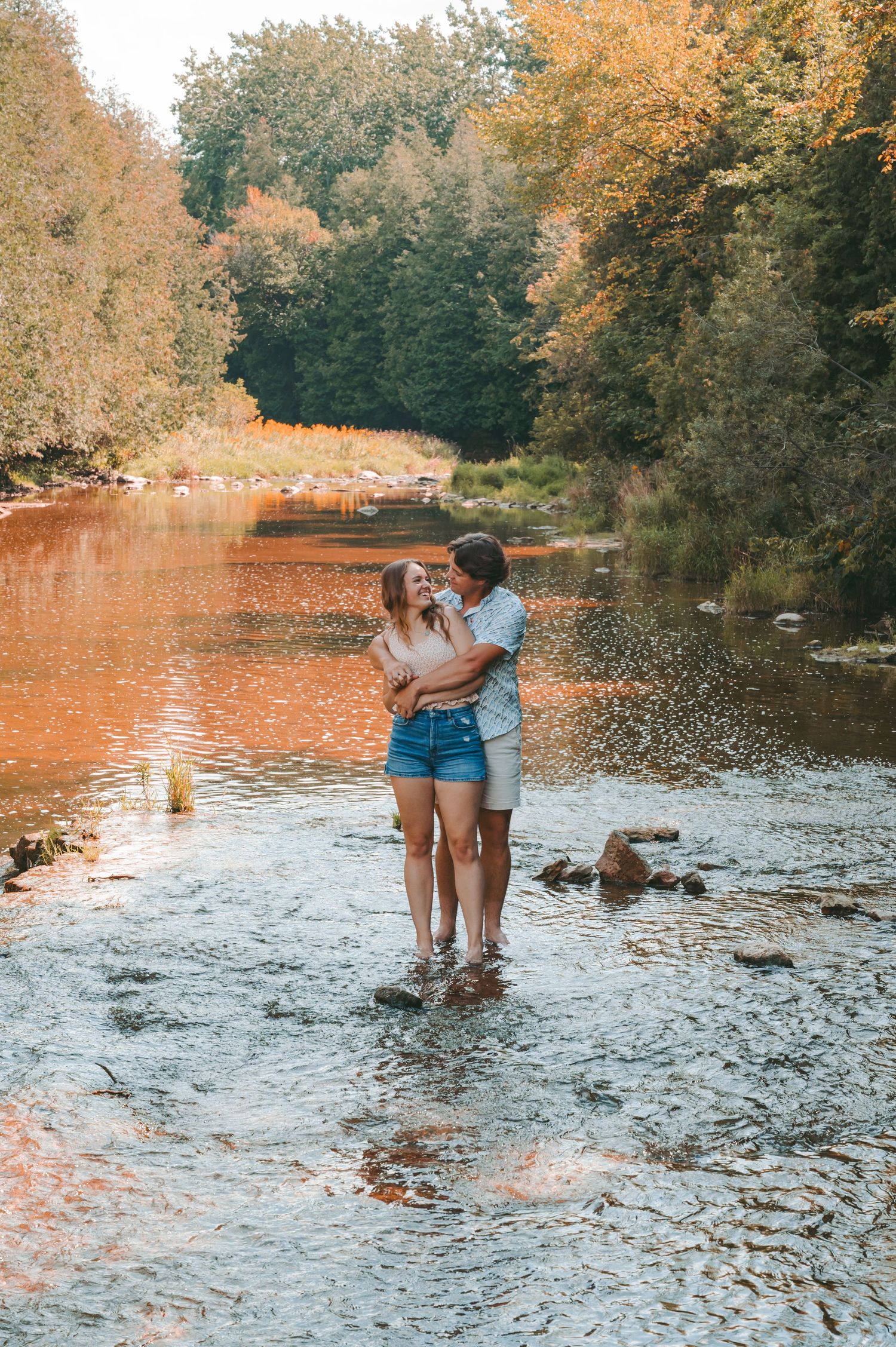 Couple sharing intimate moment while standing in shallow river during golden hour.