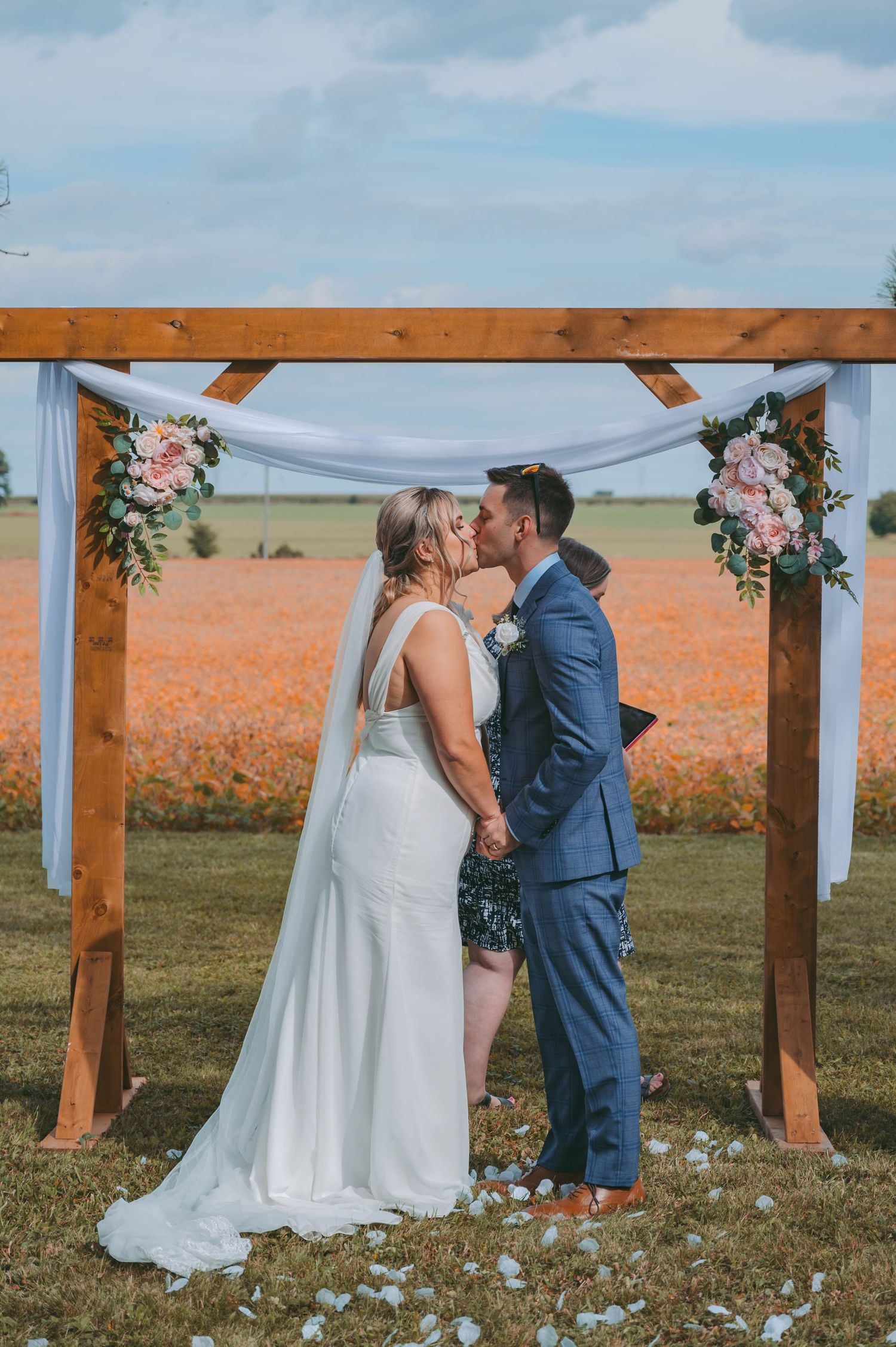 Wedding ceremony taking place under wooden pergola decorated with white fabric and flowers.