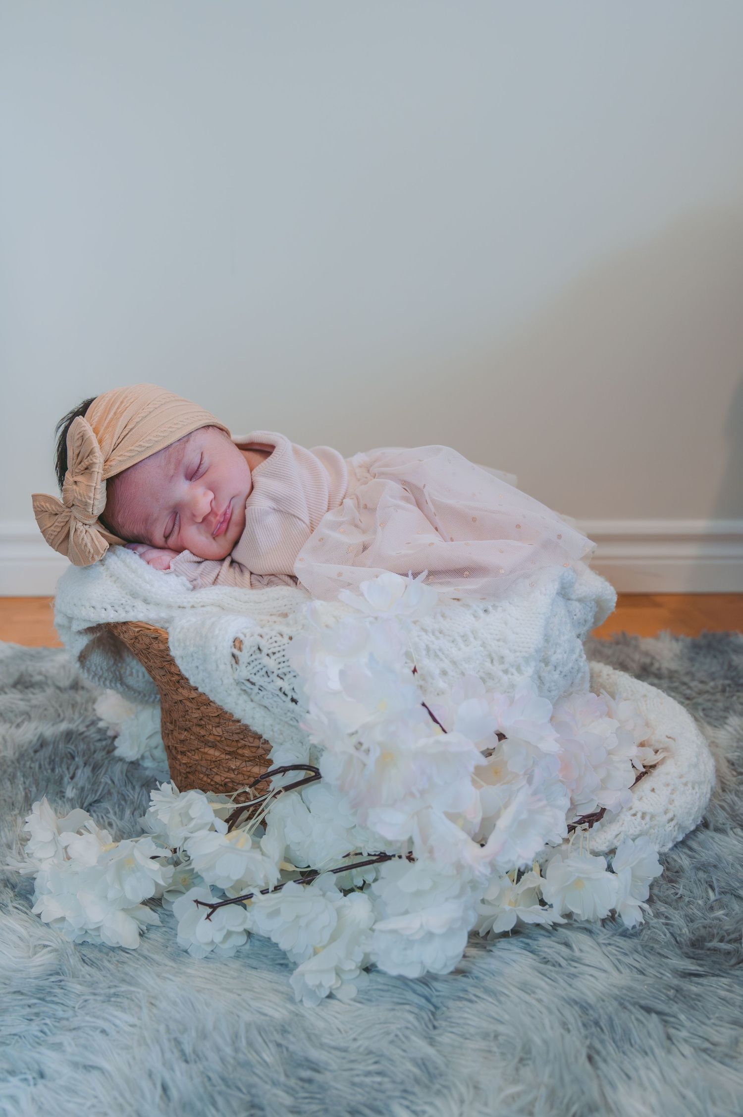 Newborn baby sleeps peacefully in basket surrounded by soft white lace and wearing bow headband.
