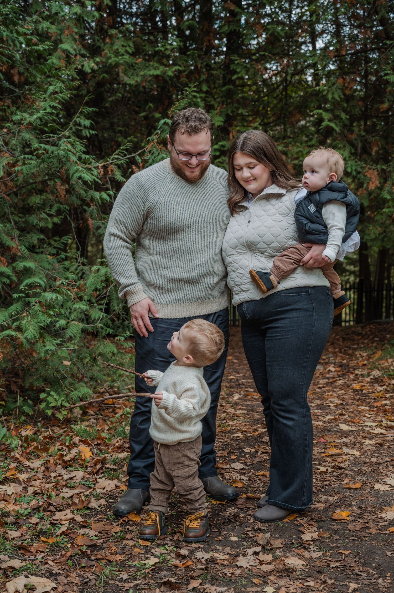 Family of four poses on a leaf-covered forest path in autumn, parents smiling at toddler holding a stick.