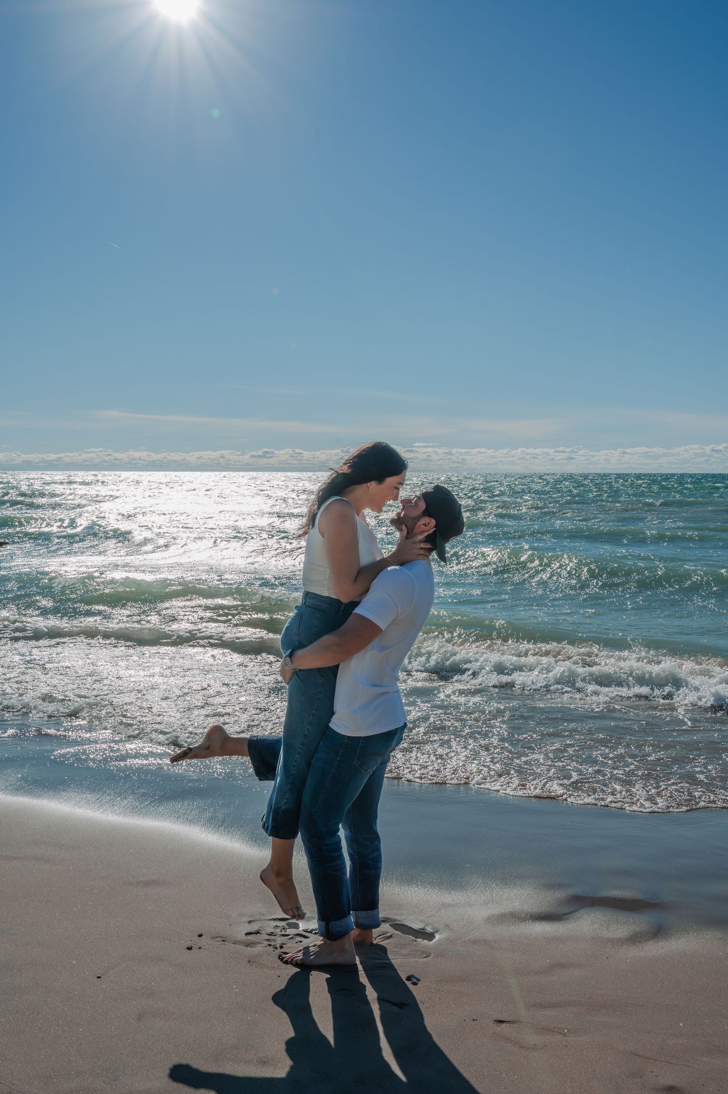 A romantic couple embraces on a sunny beach with sparkling ocean waves and bright sunlight in the background.