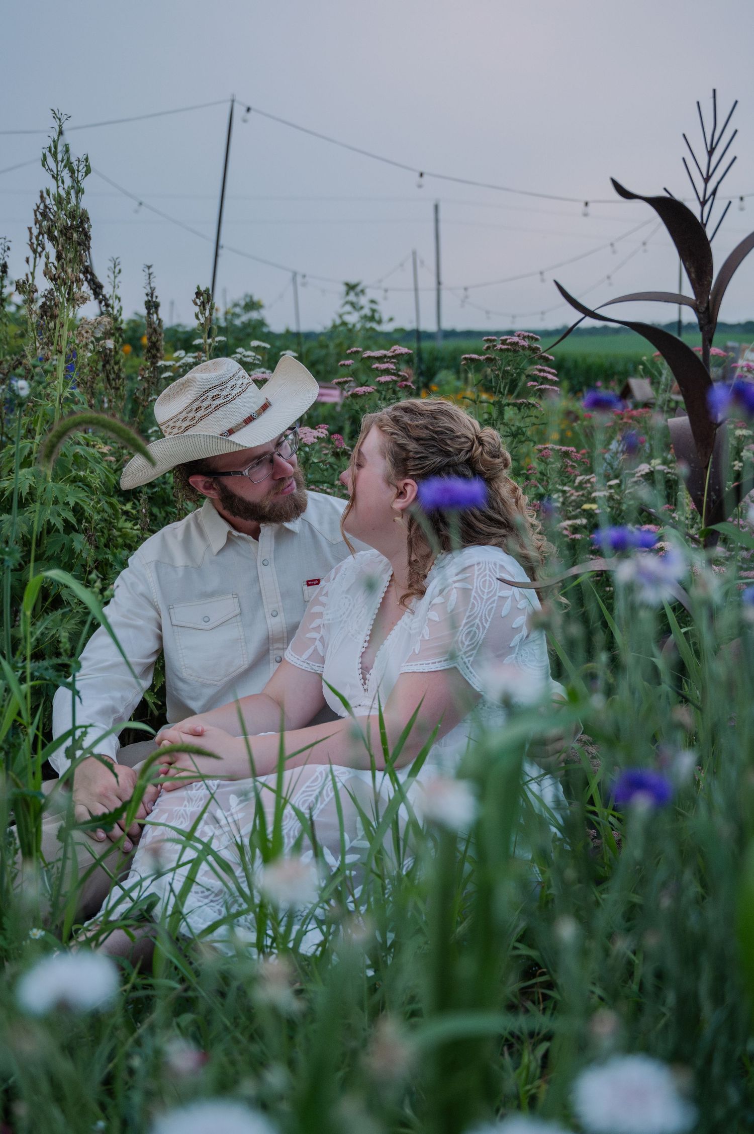 Couple sitting together in a wildflower garden, man in straw hat and woman in white dress, sharing a romantic moment.