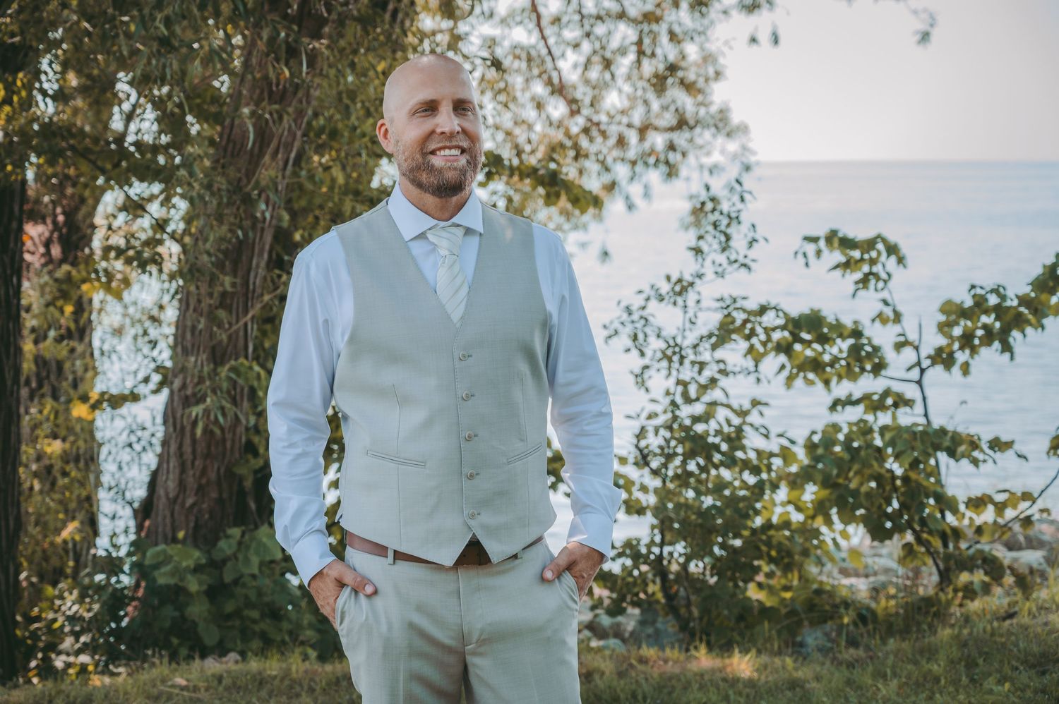 Groom in light gray vest and white shirt poses against natural waterfront backdrop.