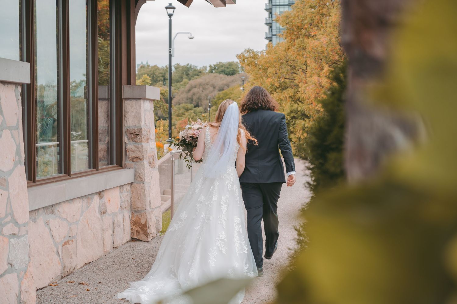 Side view of couple walking along stone building exterior in wedding attire.