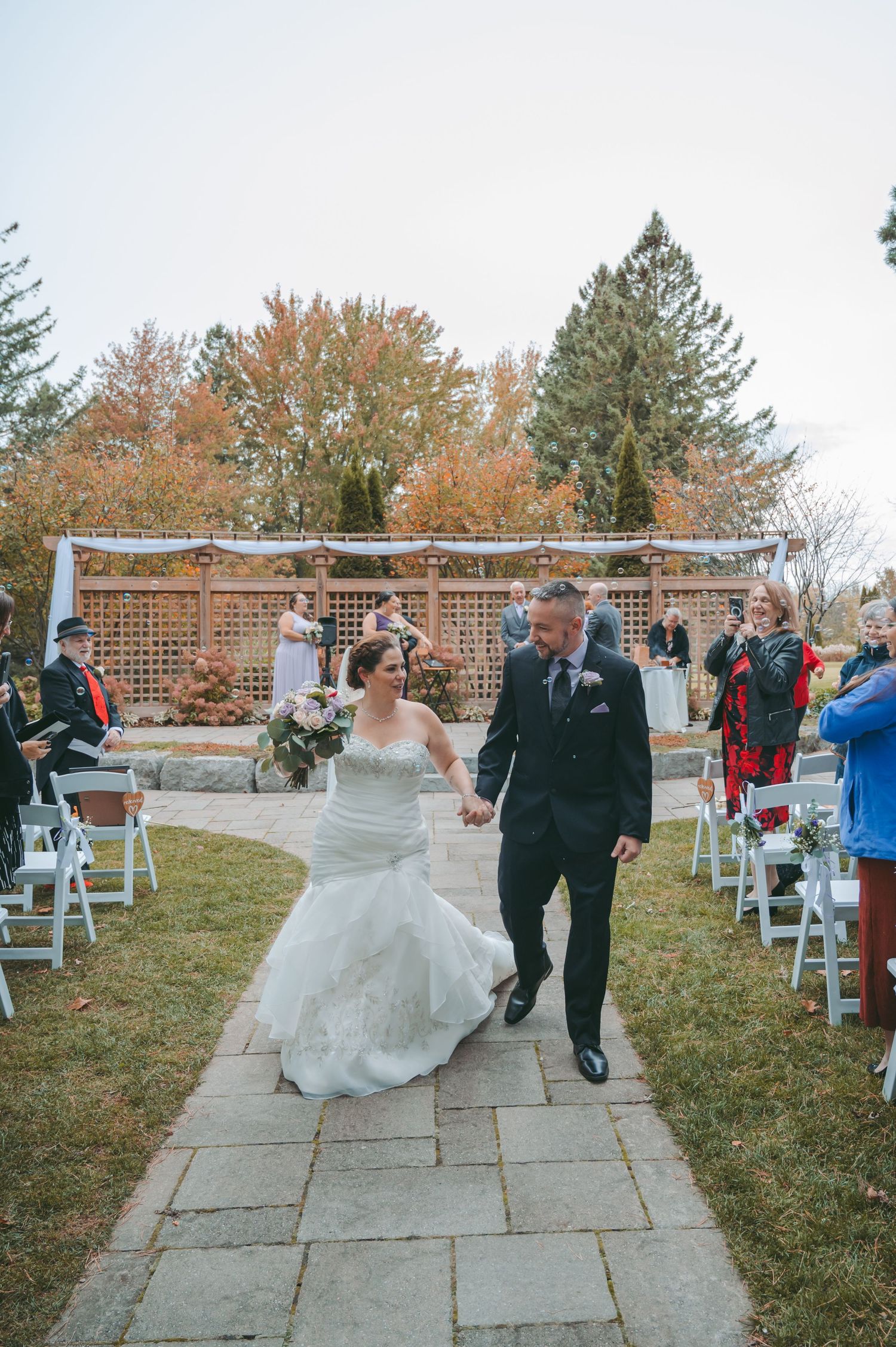 Wedding couple walks down stone path between white chairs during outdoor fall ceremony.