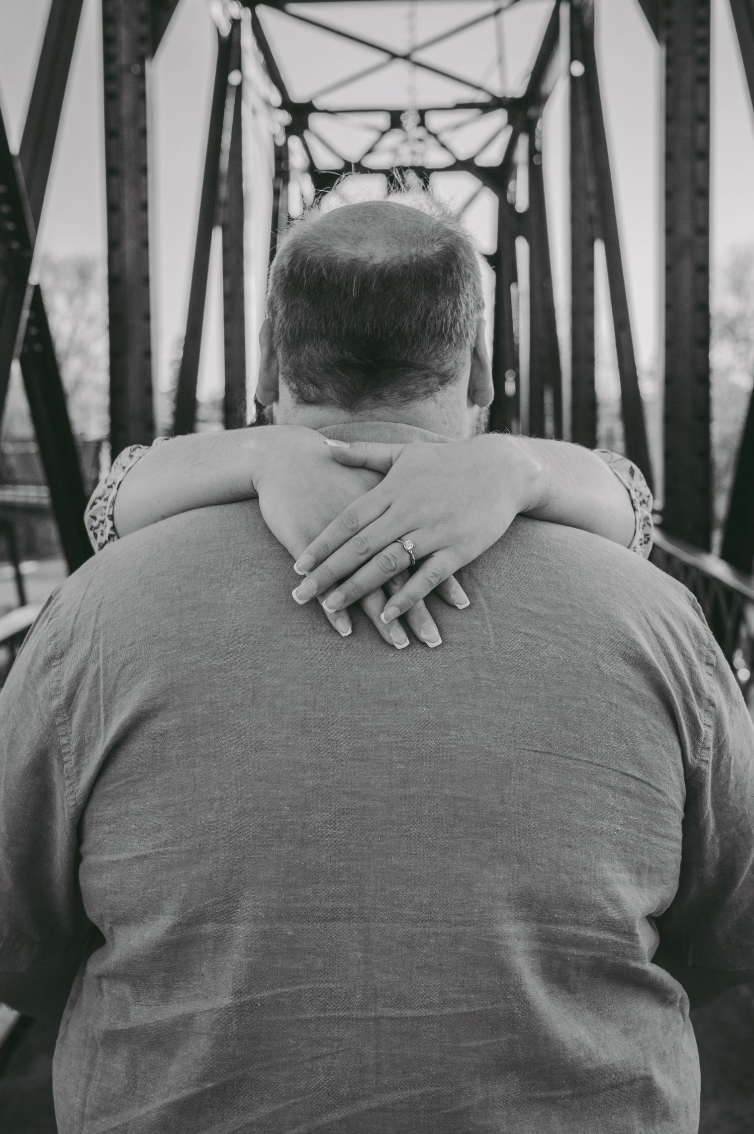 A black and white close-up photo showing hands embraced on a bridge with metal beams in the background.