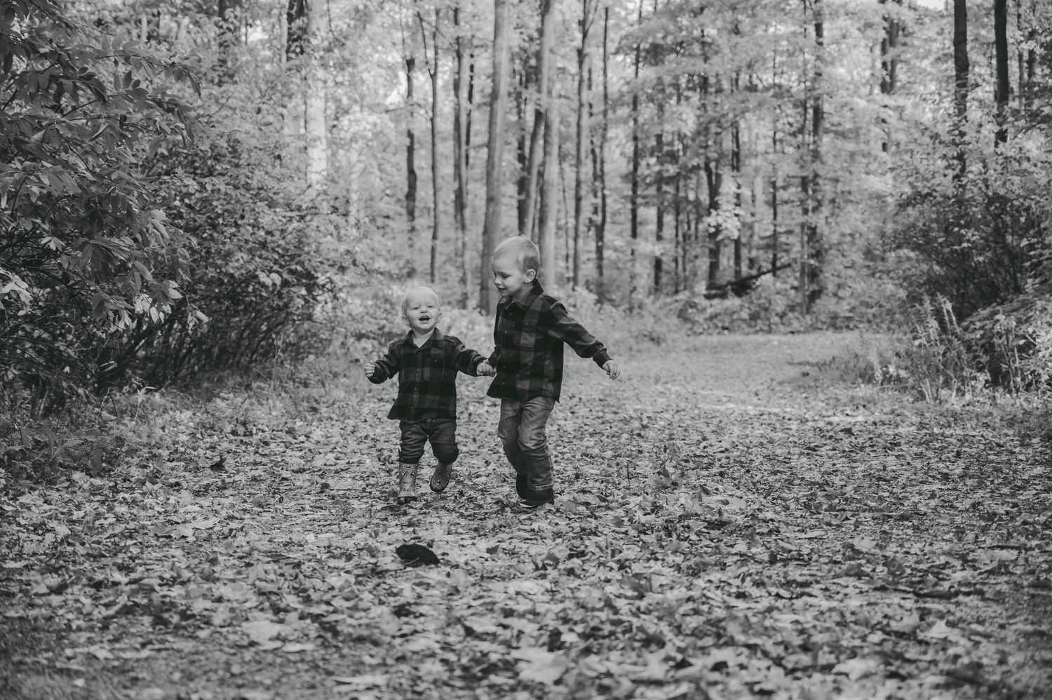 Two children running playfully through a forest path covered in fallen leaves.