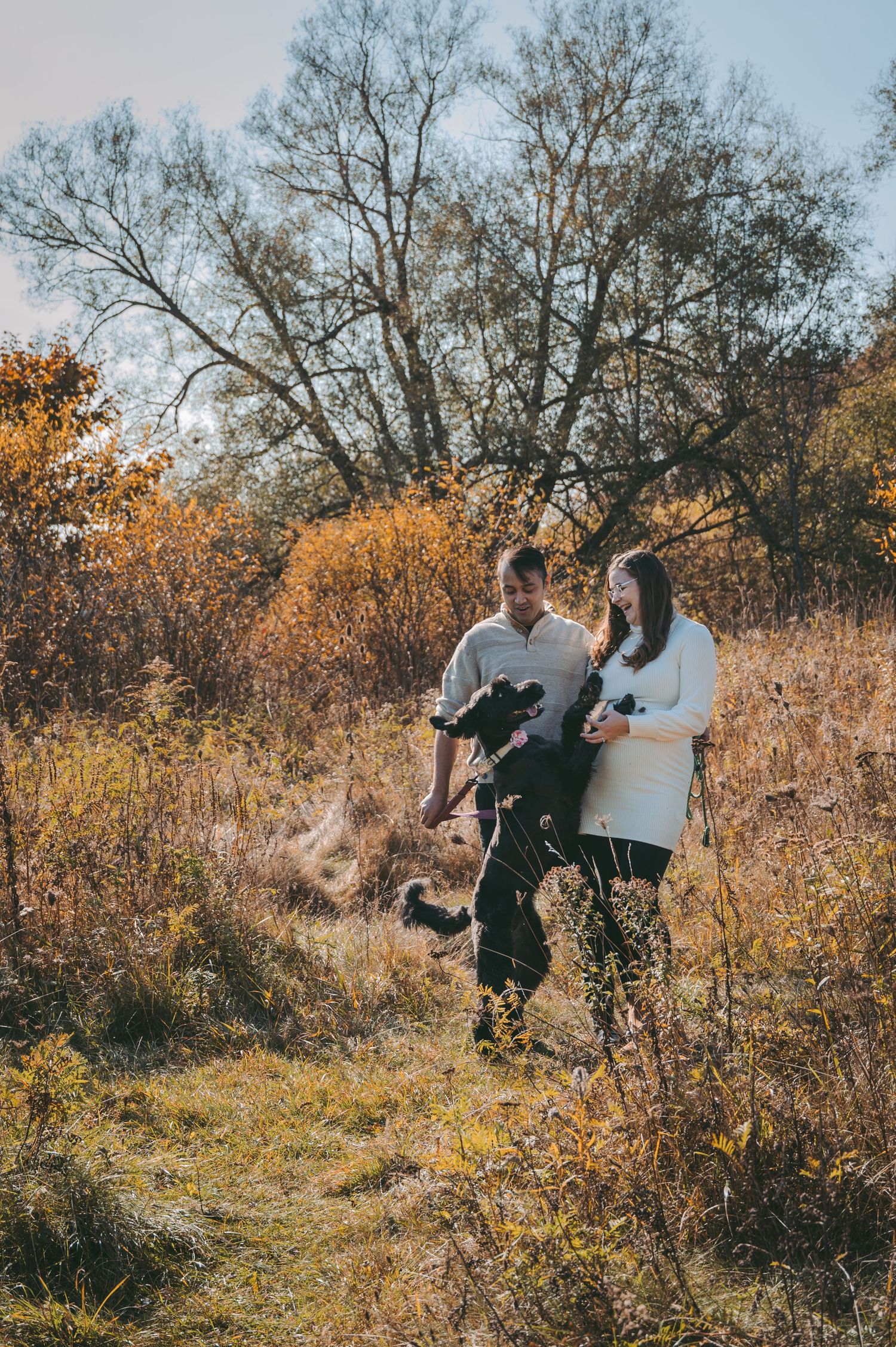 Couple walks with child through tall grass in autumn field with bare trees backdrop.