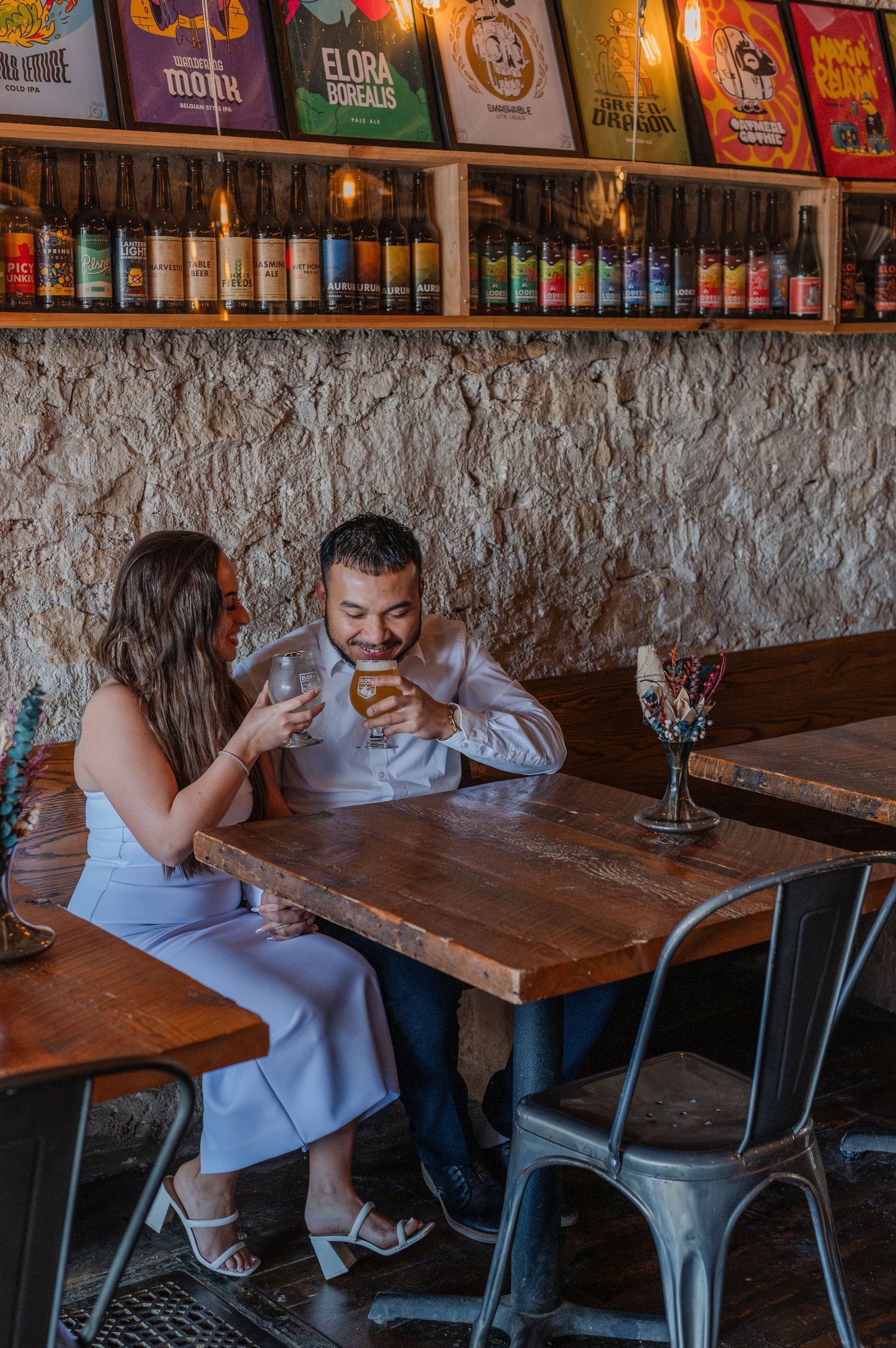 Couple sharing drinks at a rustic craft beer bar with colorful beer cans displayed on shelves.