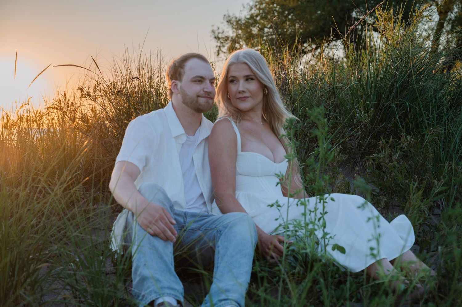 A couple sits together in a golden sunset field, the woman in white dress and man in casual attire among tall grasses.