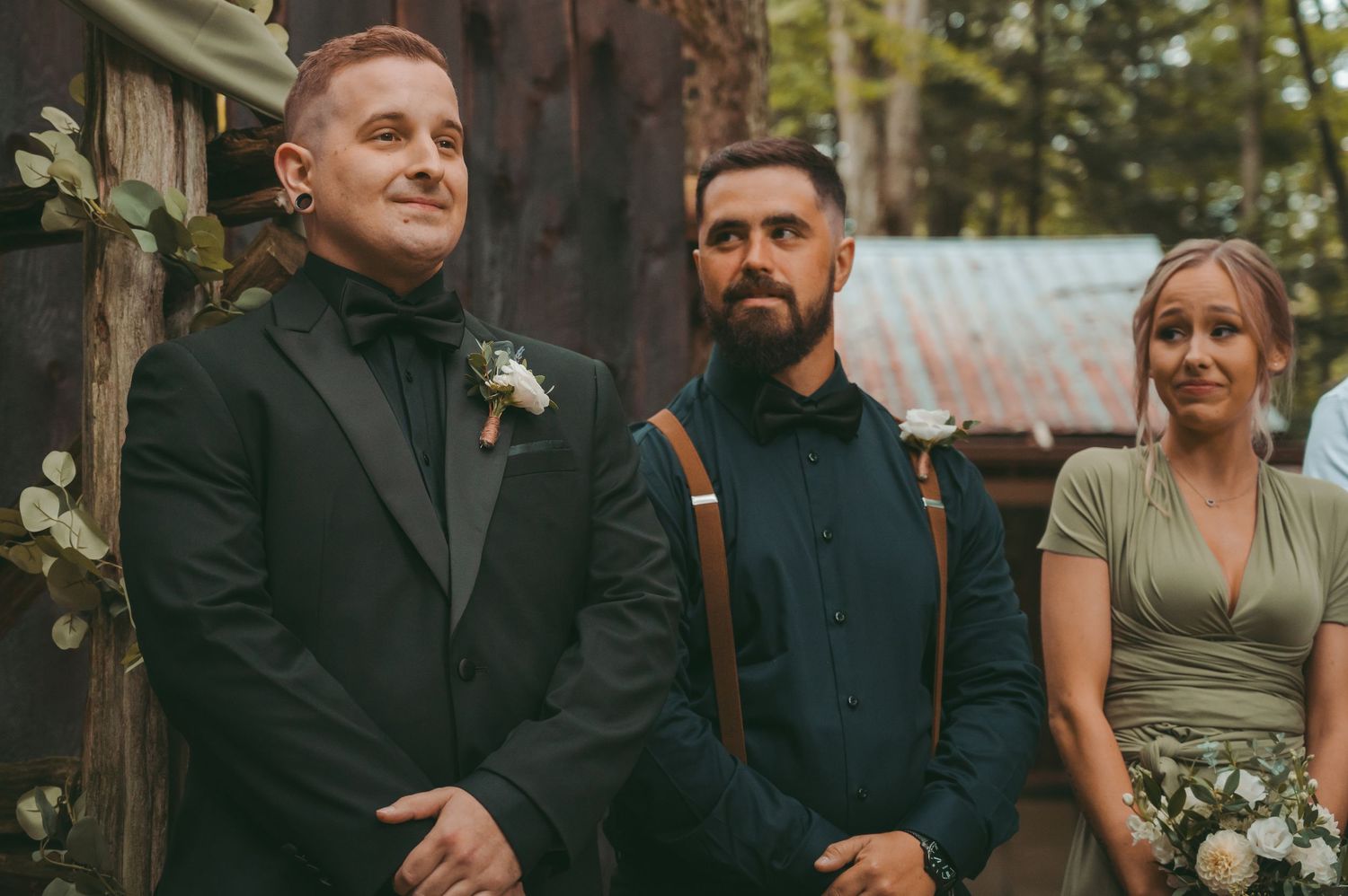 Wedding party members gather outdoors in formal attire with greenery and rustic wooden elements in the background.