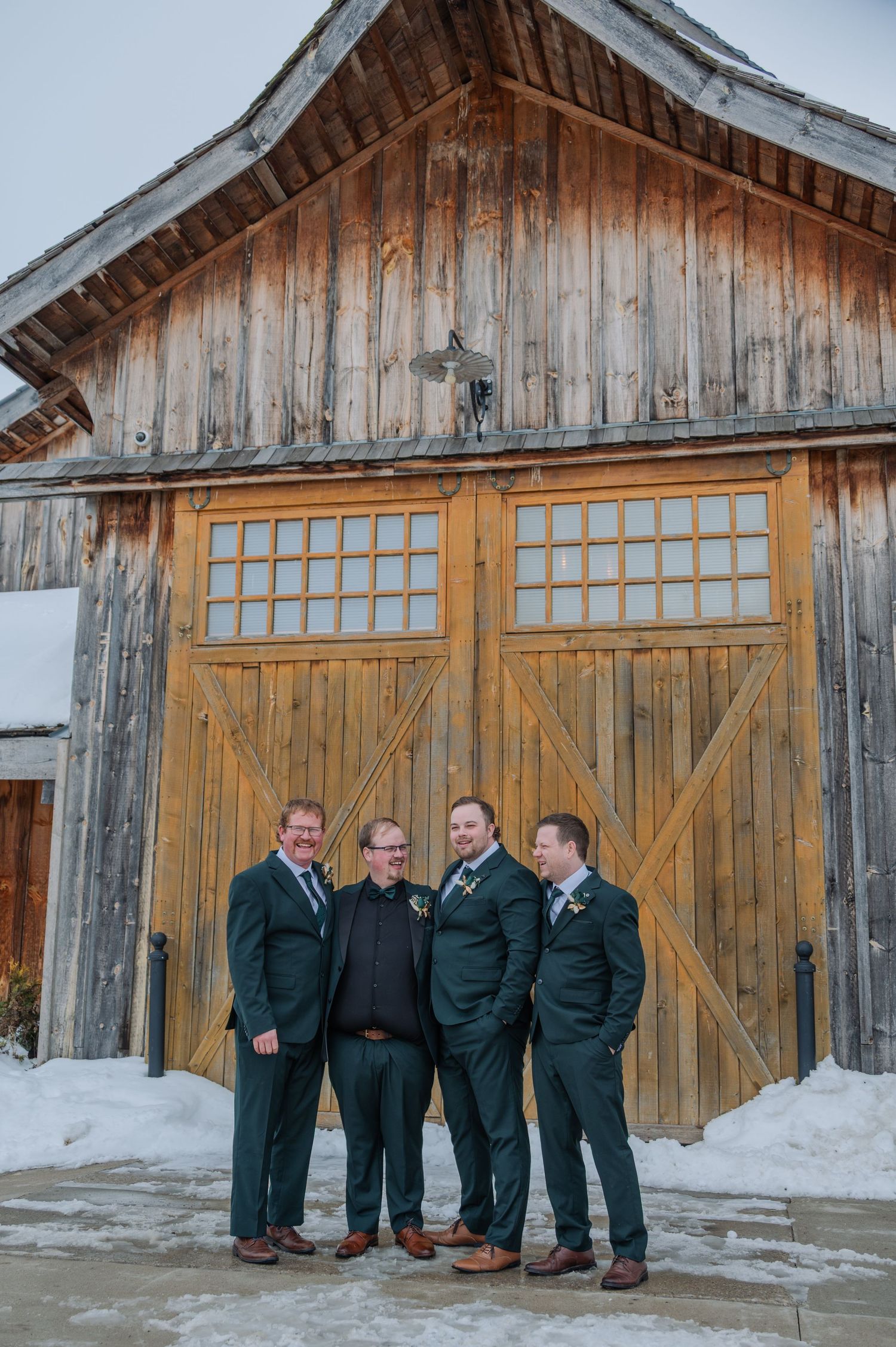 Four groomsmen in dark green suits pose together in front of a rustic wooden barn in a snowy winter setting.