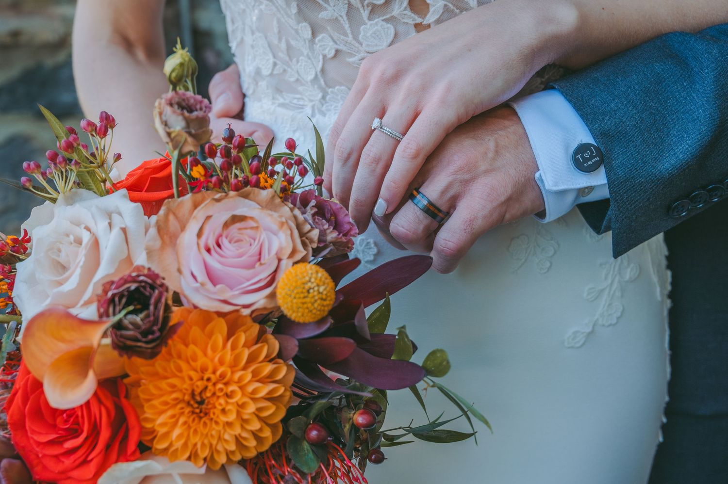 Close-up detail of hands, rings, and vibrant autumn wedding bouquet with orange and pink flowers.