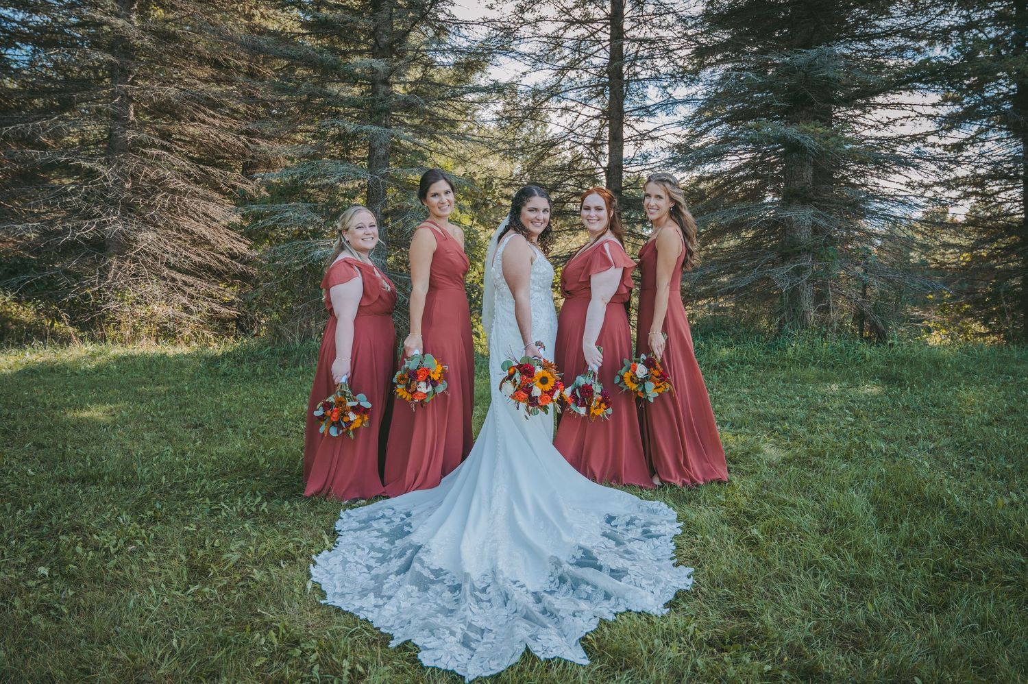 Bride in lace gown poses with bridesmaids in burgundy dresses holding autumn-colored bouquets.