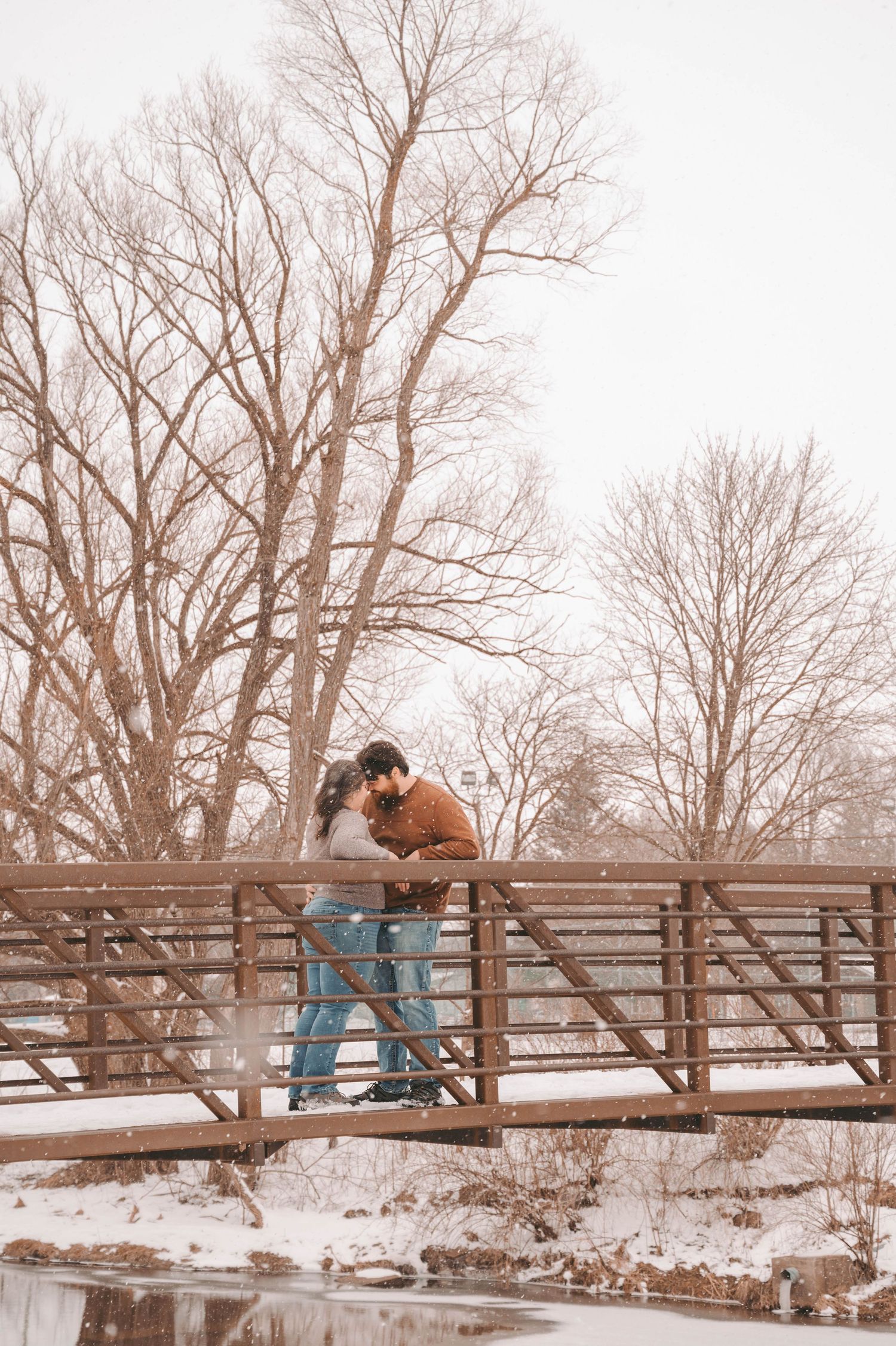 A couple embracing on a wooden bridge during winter with bare trees in the background.