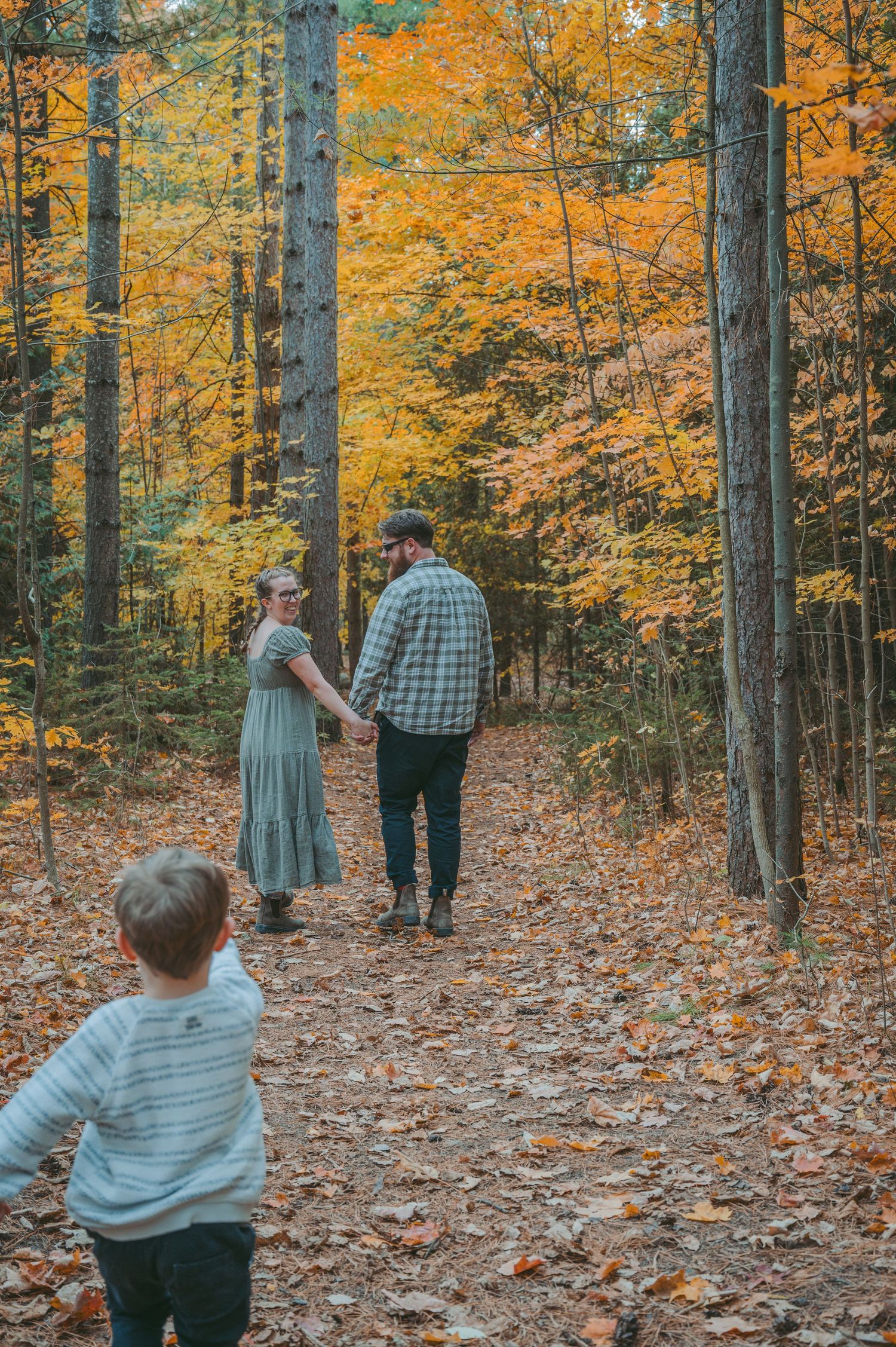 Child follows parents walking hand in hand on leaf-covered autumn forest path.