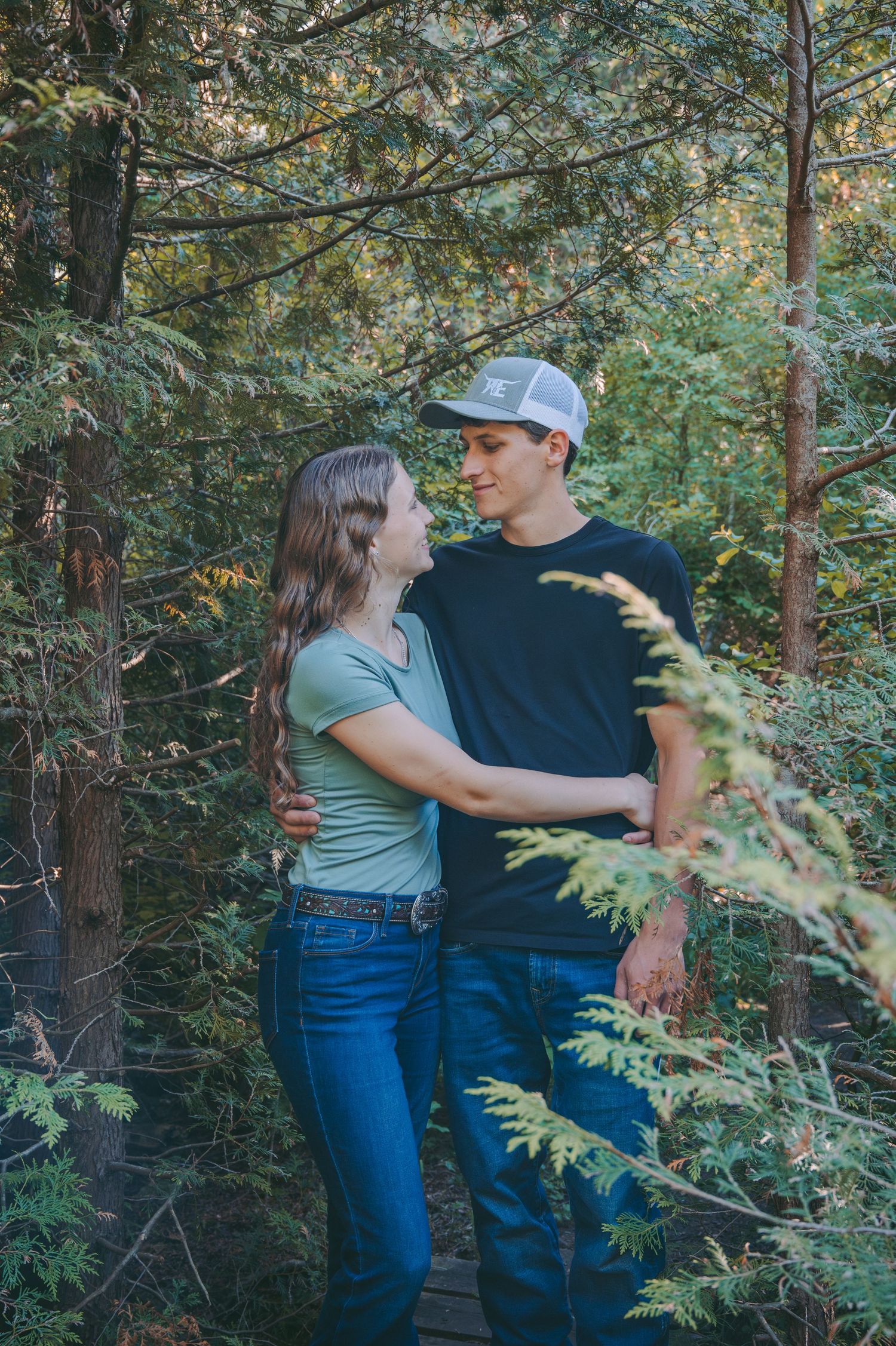 A romantic couple embraces in a wooded forest setting with dappled sunlight filtering through the trees.