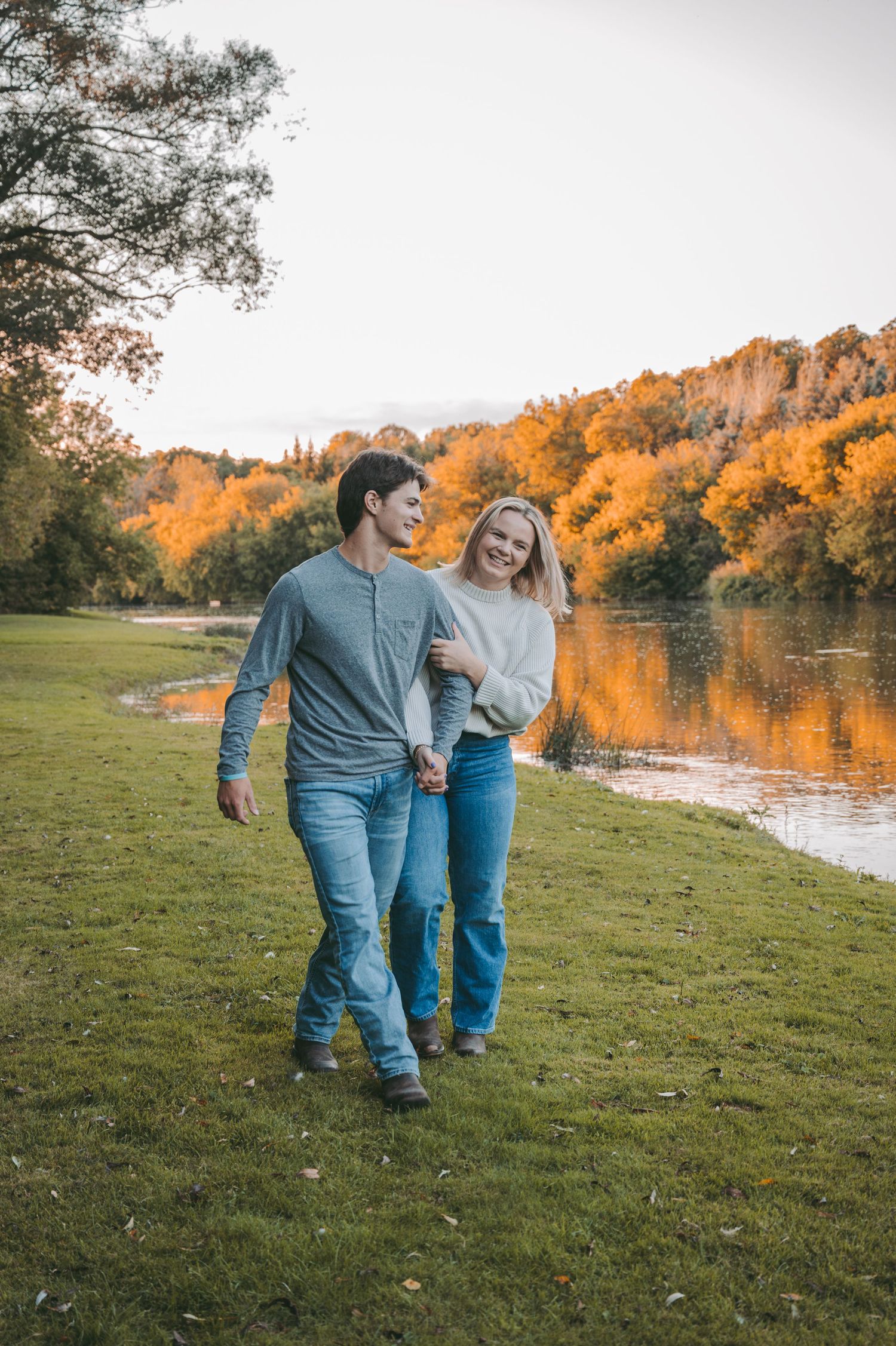 A couple strolls together along a riverbank with autumn foliage reflecting in the water.
