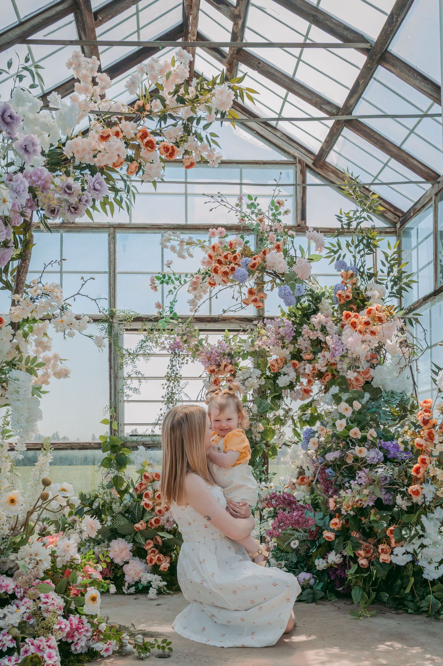 Intimate mother-child moment in sunlit greenhouse surrounded by cascading floral arrangements.