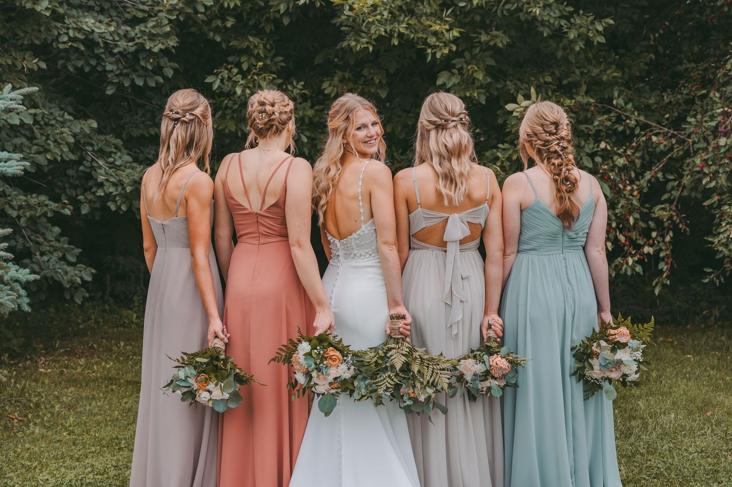 Back view of bridal party in pastel dresses holding bouquets on green lawn.
