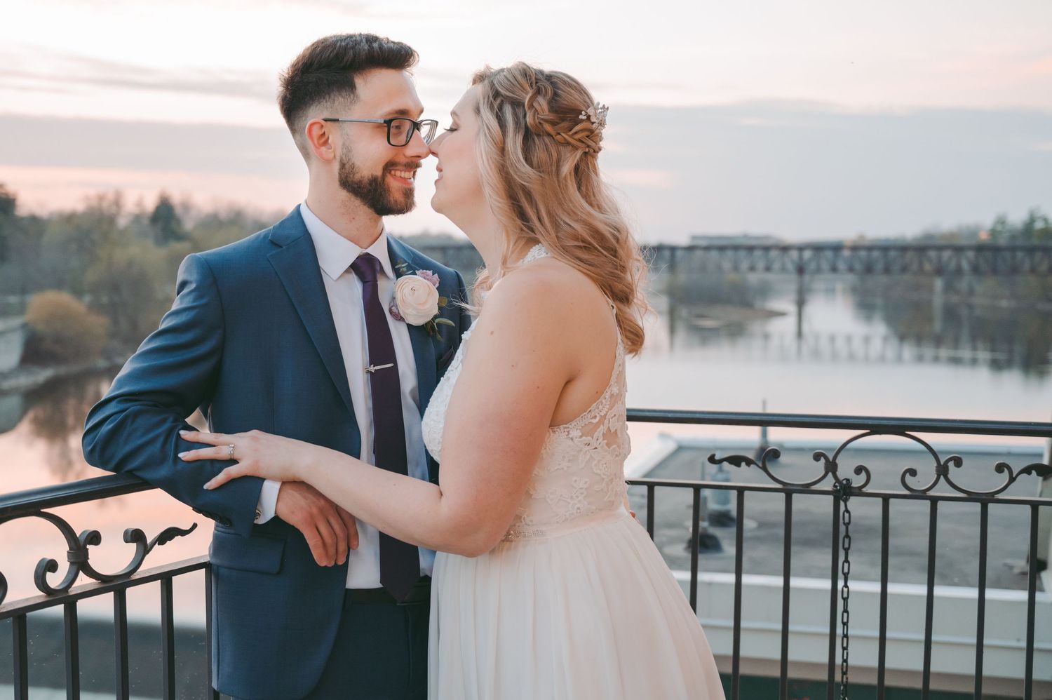 Wedding couple sharing intimate moment on balcony overlooking bridge at sunset.
