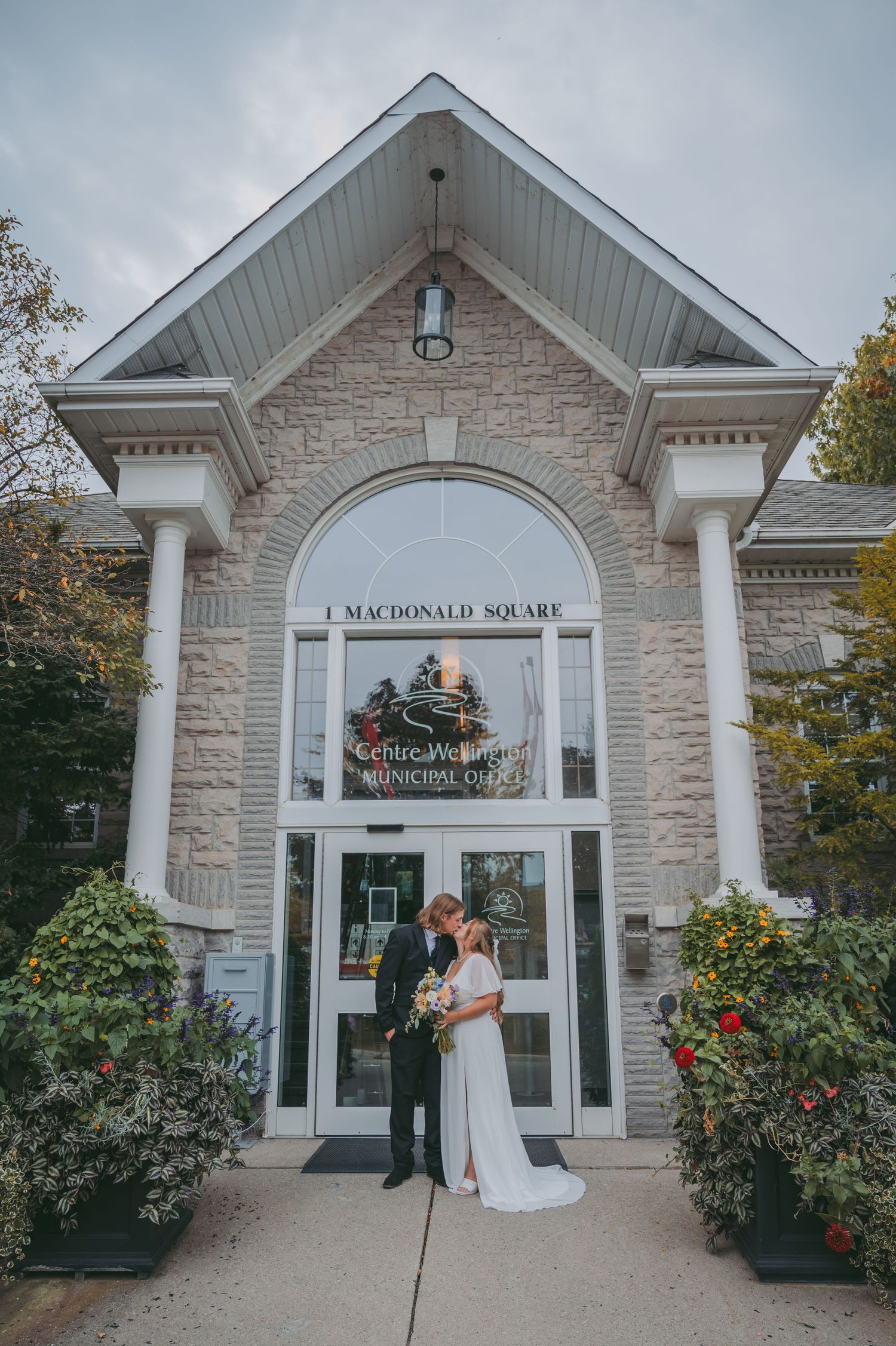A stone building entrance with white columns and arched windows showcasing a wedding couple in front of Macdonald Square, Elora.