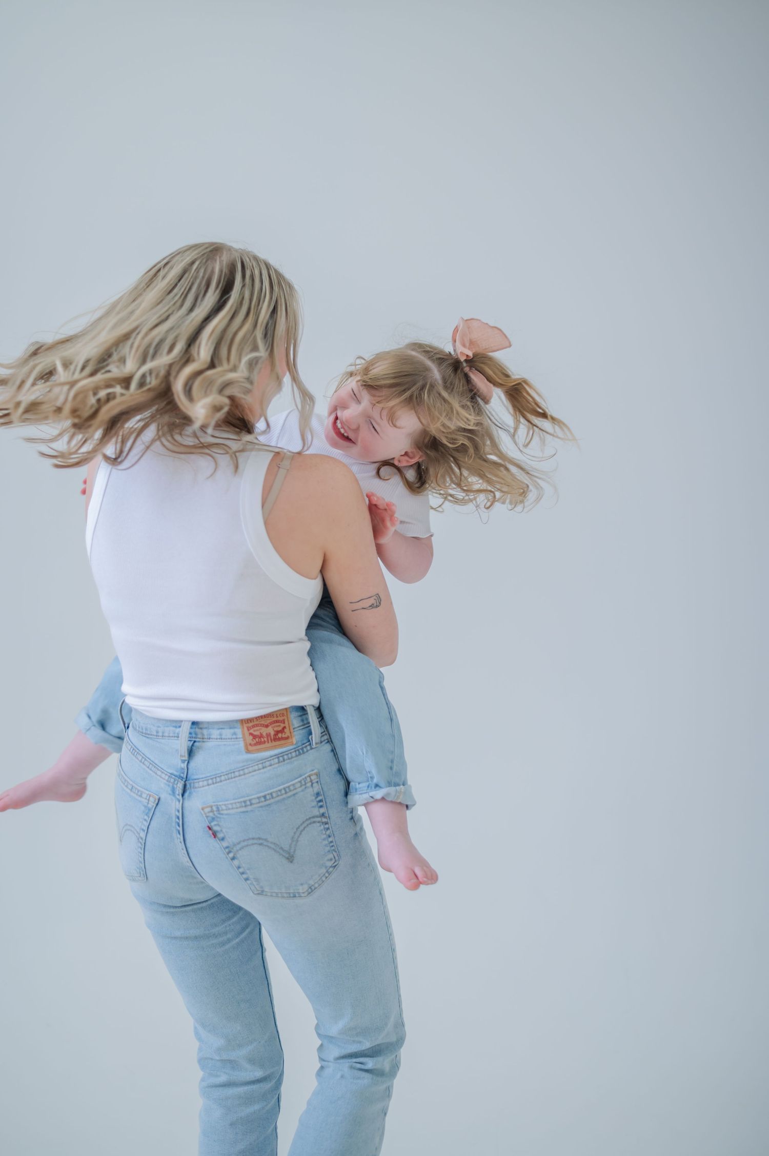 A mother in Levi jeans and white top joyfully spins her laughing blonde toddler daughter in a bright studio.