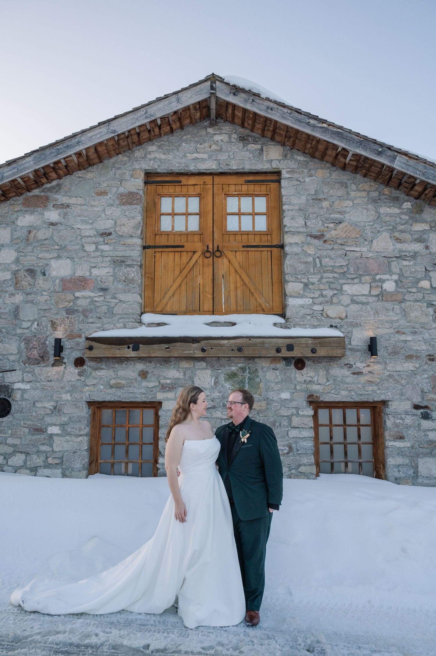 Bride and groom stand together in snow before a rustic stone barn with wooden doors during a winter wedding.