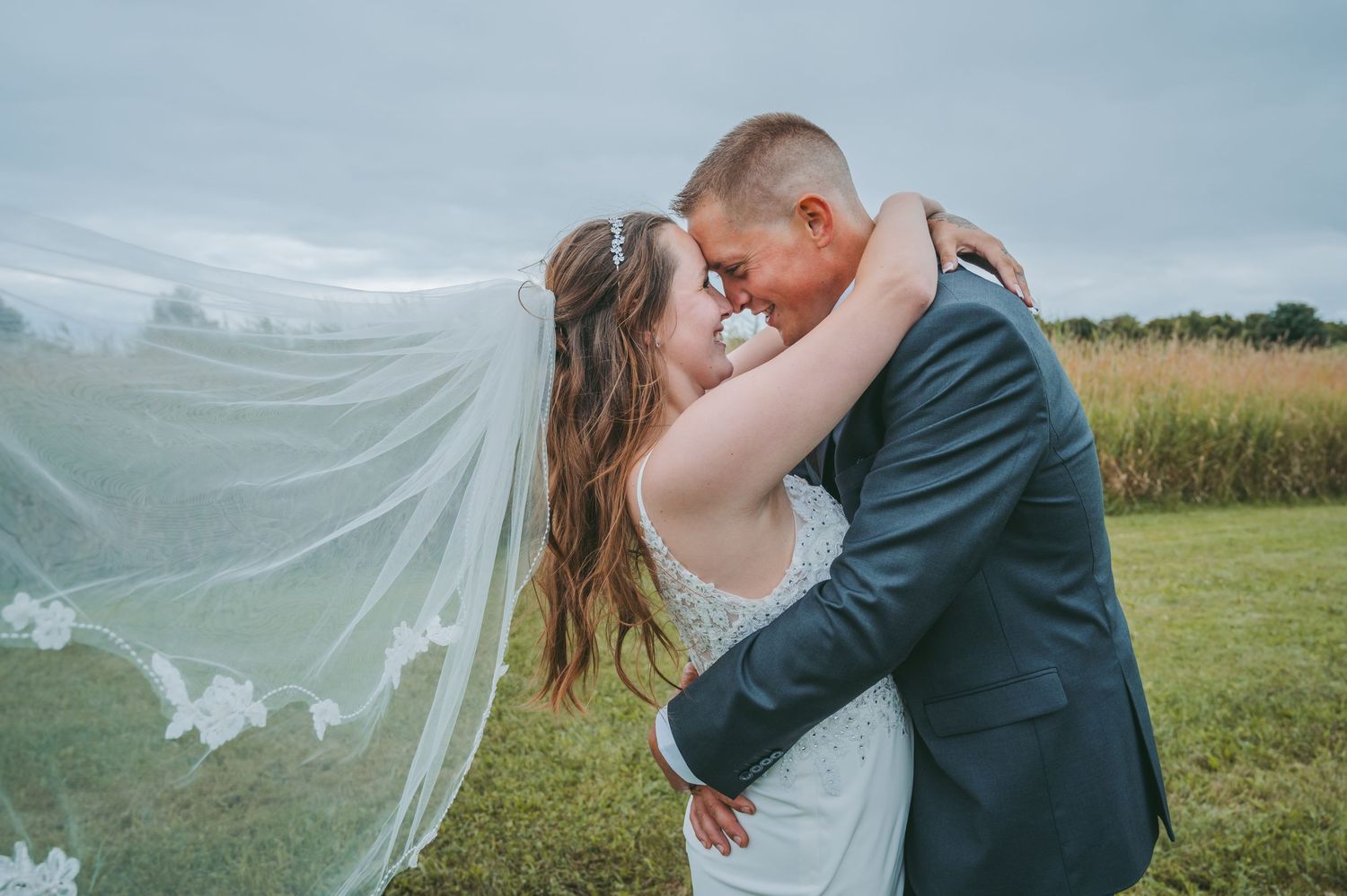 Bride and groom share a romantic kiss outdoors as her lace veil billows in the wind on their wedding day.
