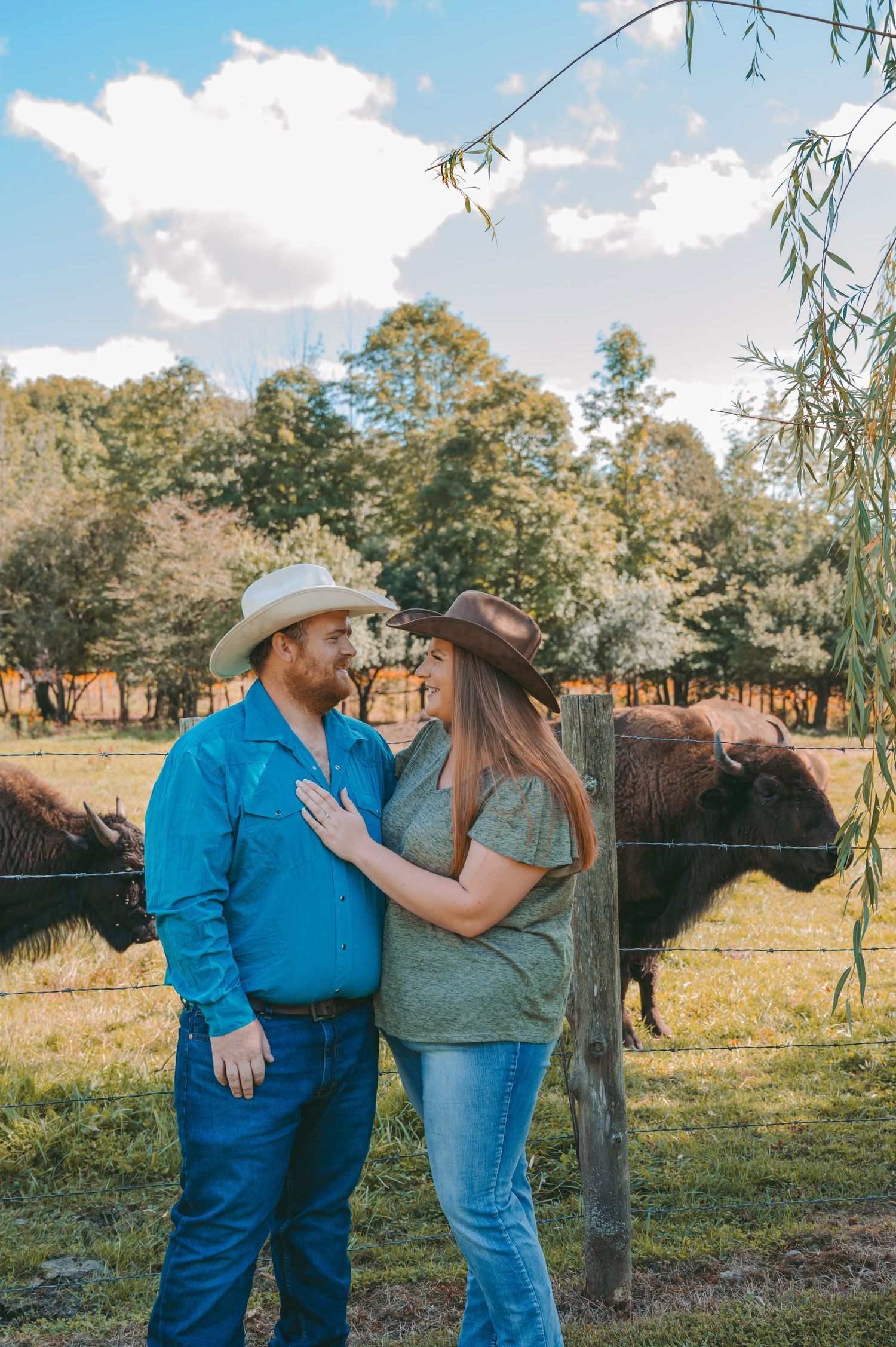 A couple in country western attire share a tender moment near a fence with cattle in the background.