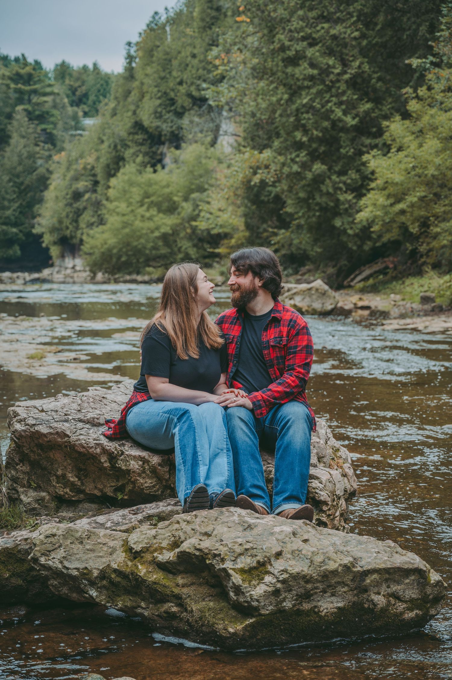 Couple sitting close together on large rock overlooking scenic river and forest landscape.