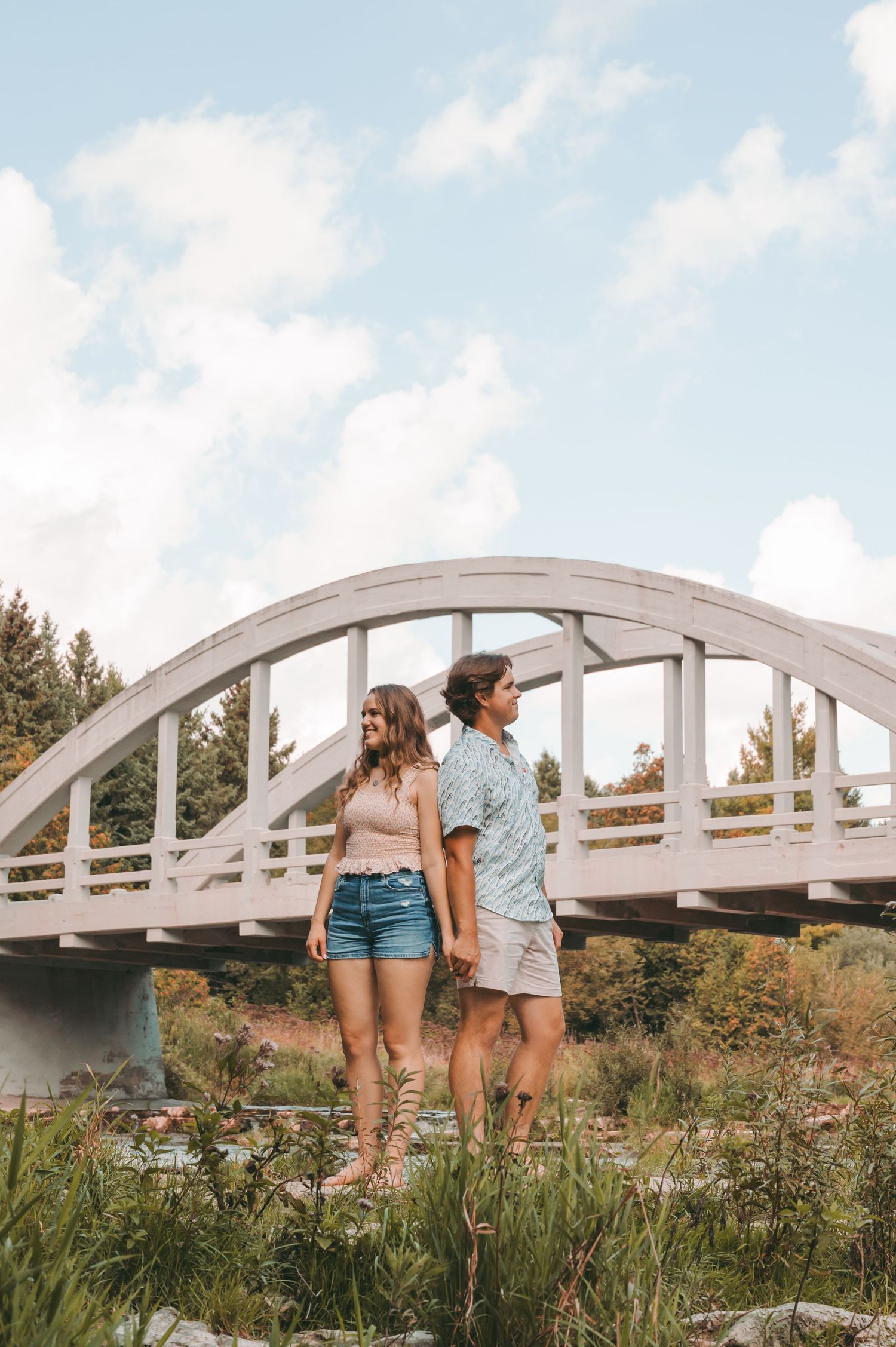Couple standing by white arched bridge on sunny day surrounded by nature and greenery.