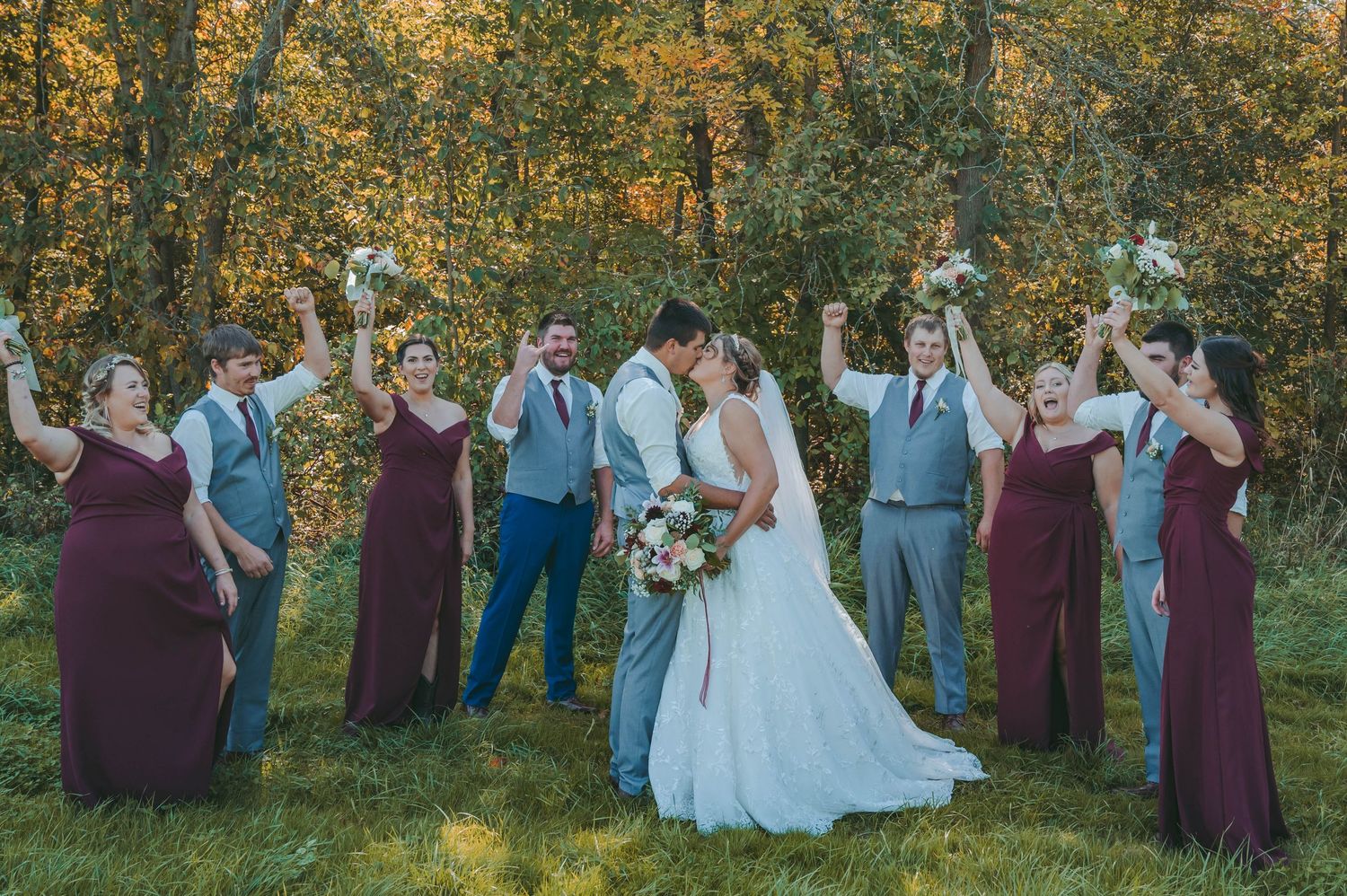 Wedding party in burgundy and blue attire poses together outdoors with autumn foliage in background.