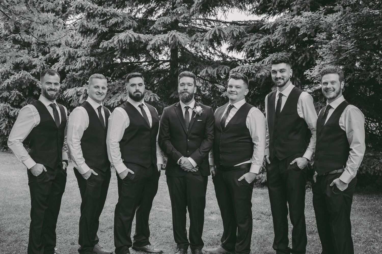 A group of groomsmen in black vests and white shirts pose together for a formal wedding photo outdoors.