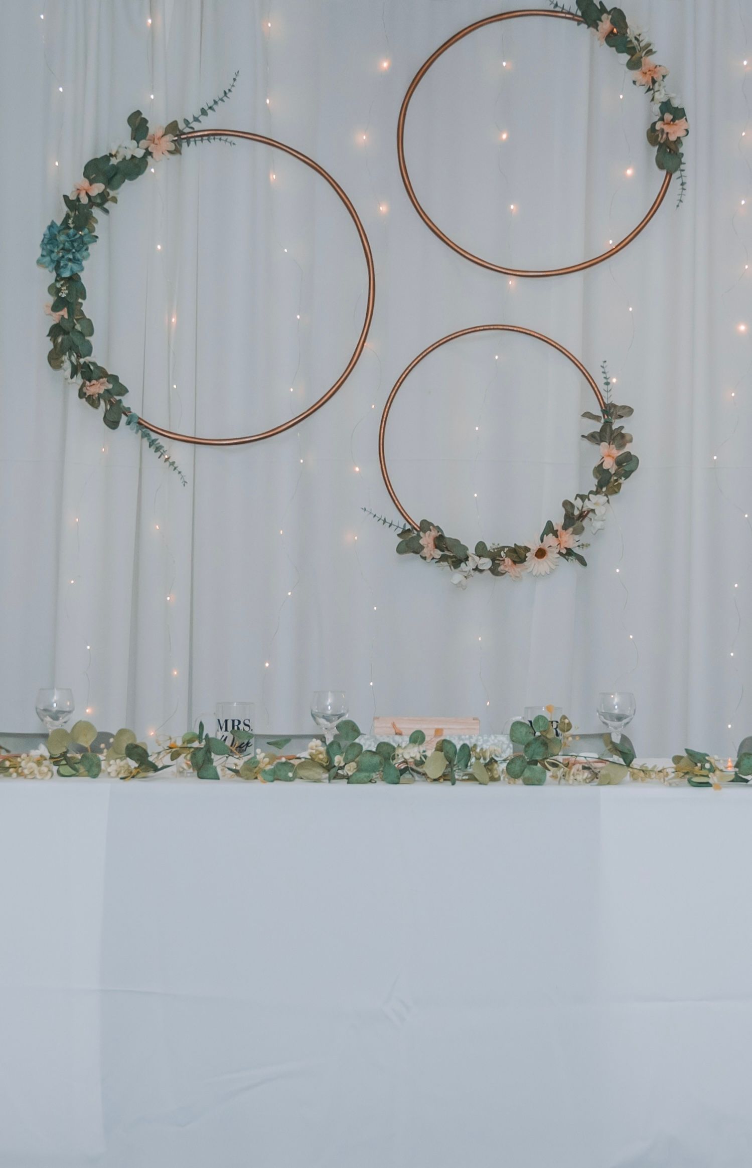 Three decorative gold hoops adorned with eucalyptus and flowers hang against a white draped backdrop with twinkle lights.