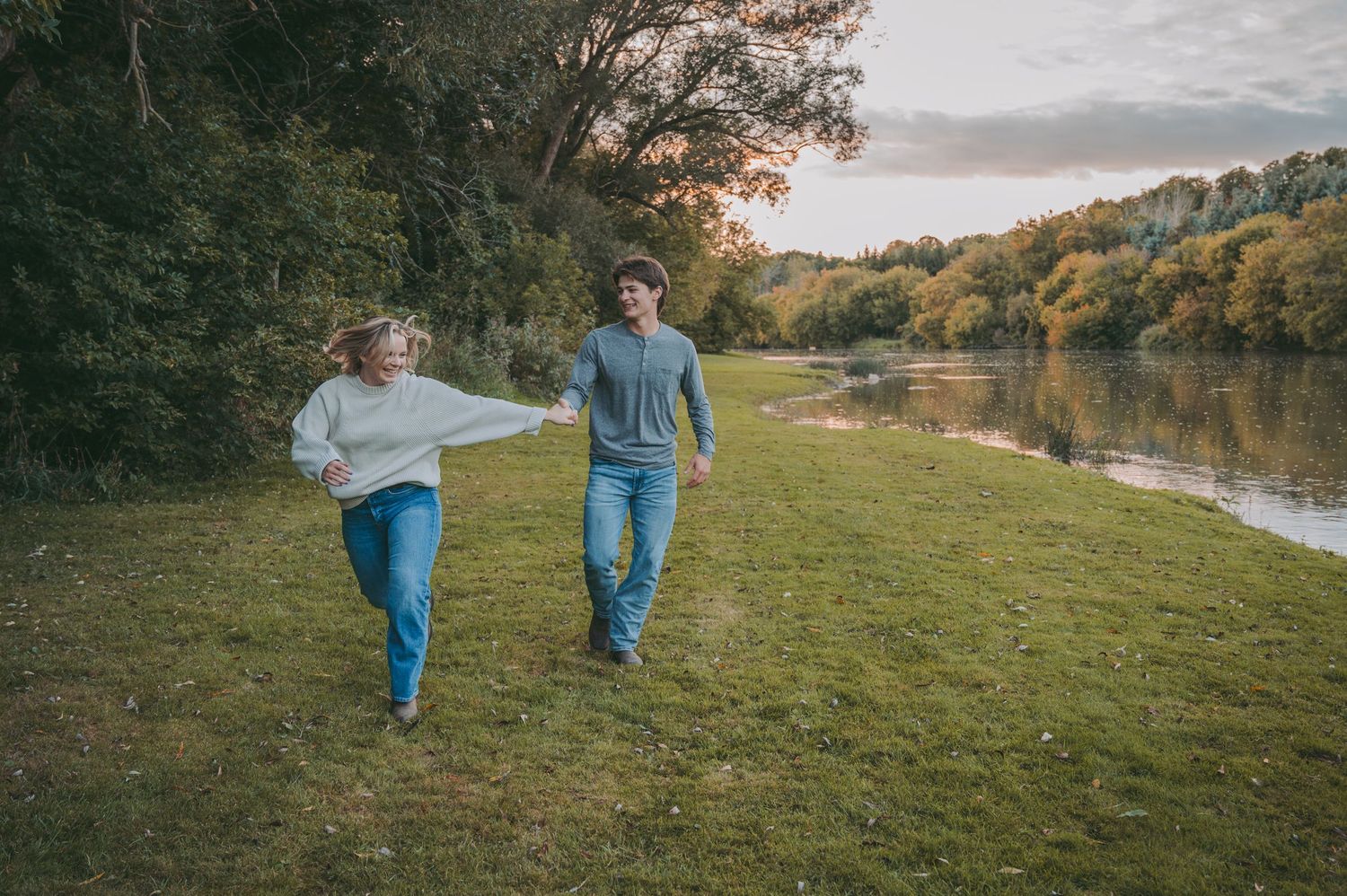A playful couple running together on grass alongside a peaceful lake.