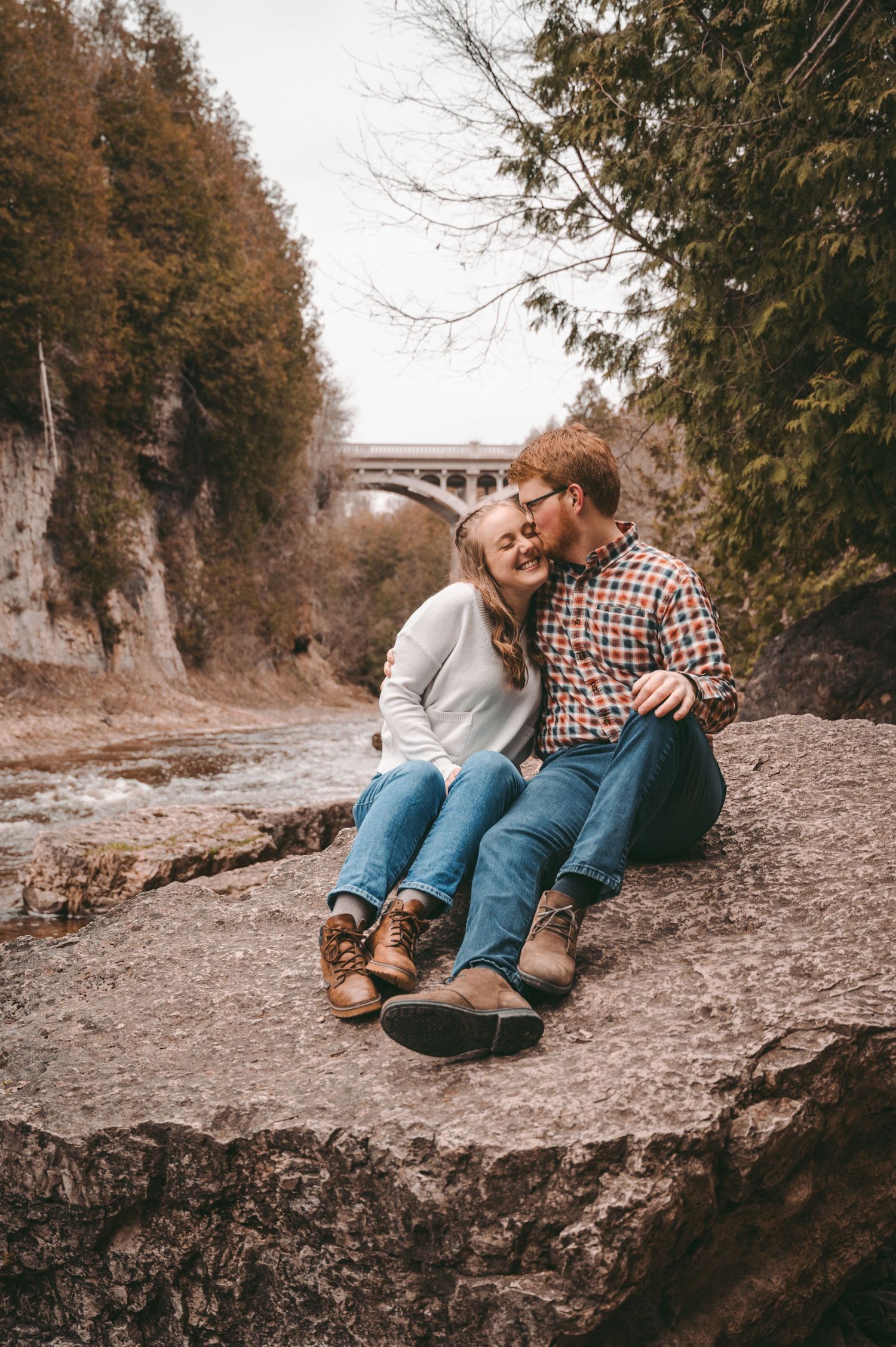 Couple sits together on rocky outcrop with bridge visible through autumn trees in background.