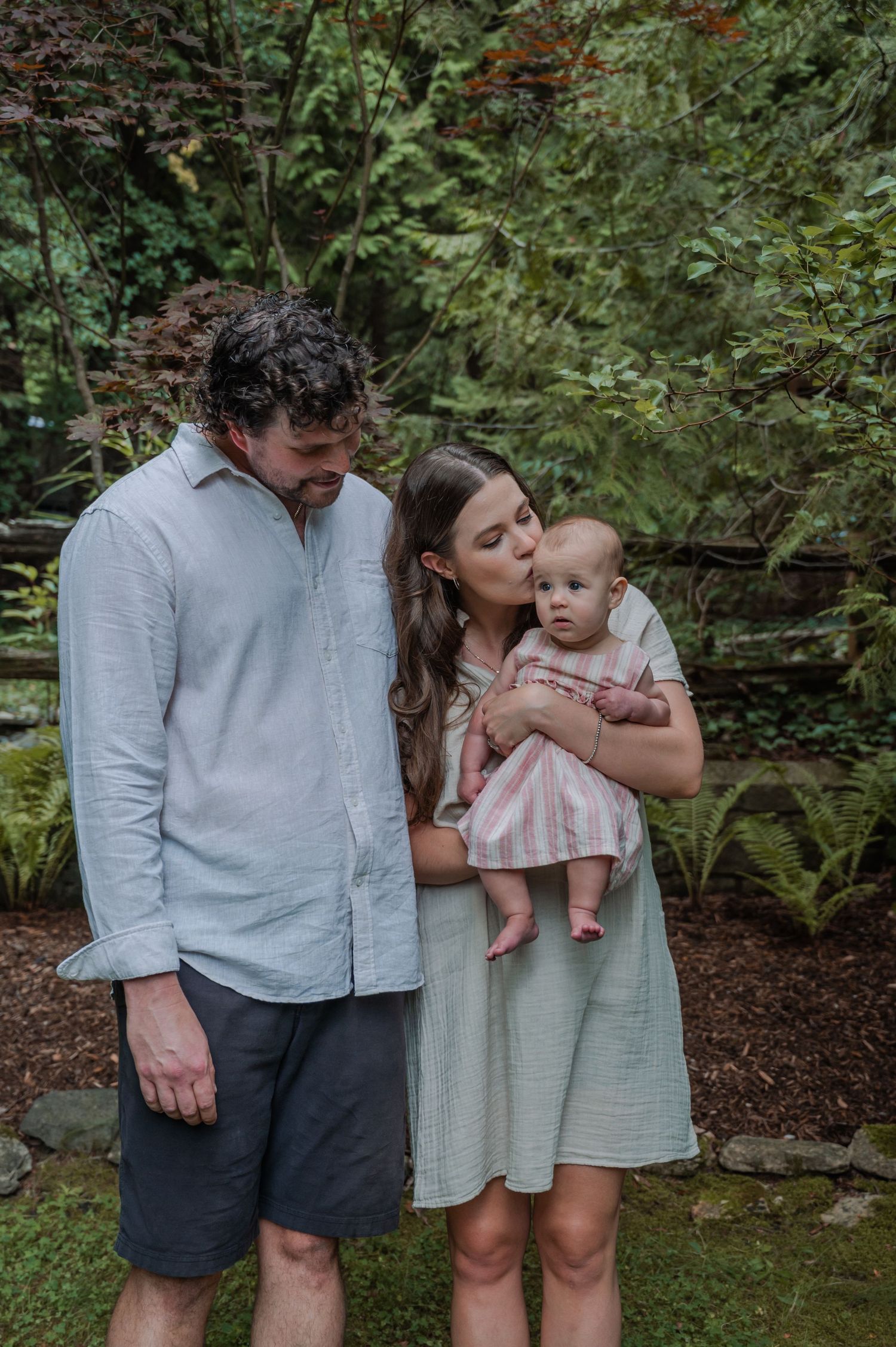 In a lush garden setting, a family shares tender moments with their baby, wearing coordinated light-colored outfits.
