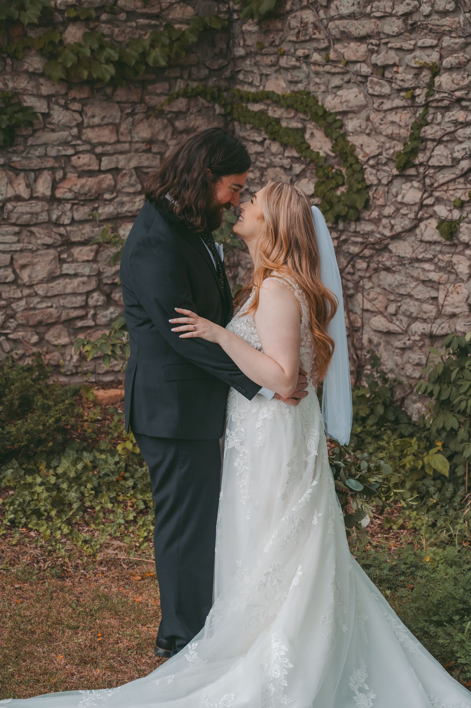 A bride and groom share a romantic moment in front of a stone wall covered in ivy, smiling at each other.