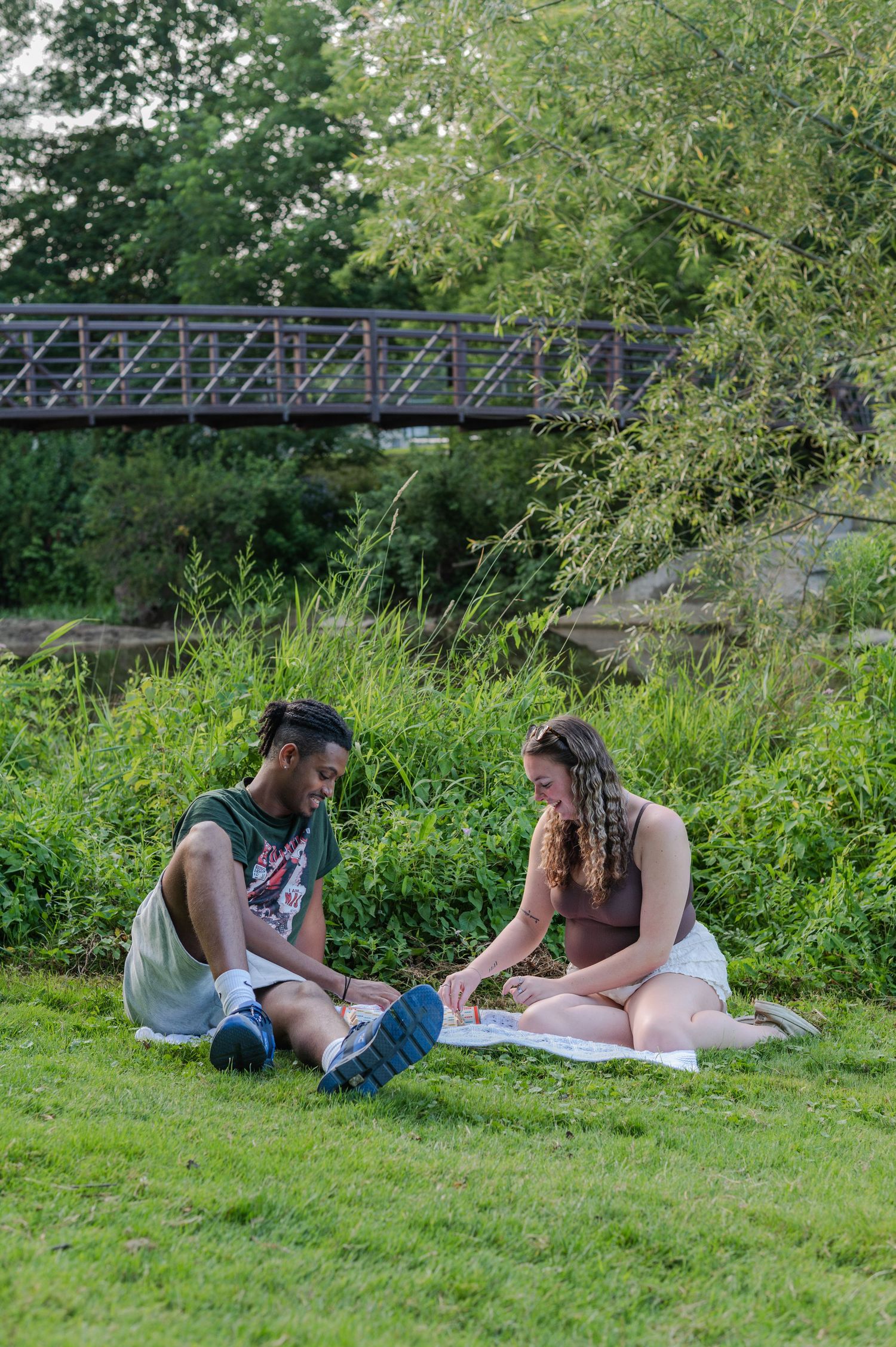 Couple sitting on grass blanket in park near bridge, enjoying outdoor picnic on sunny day.